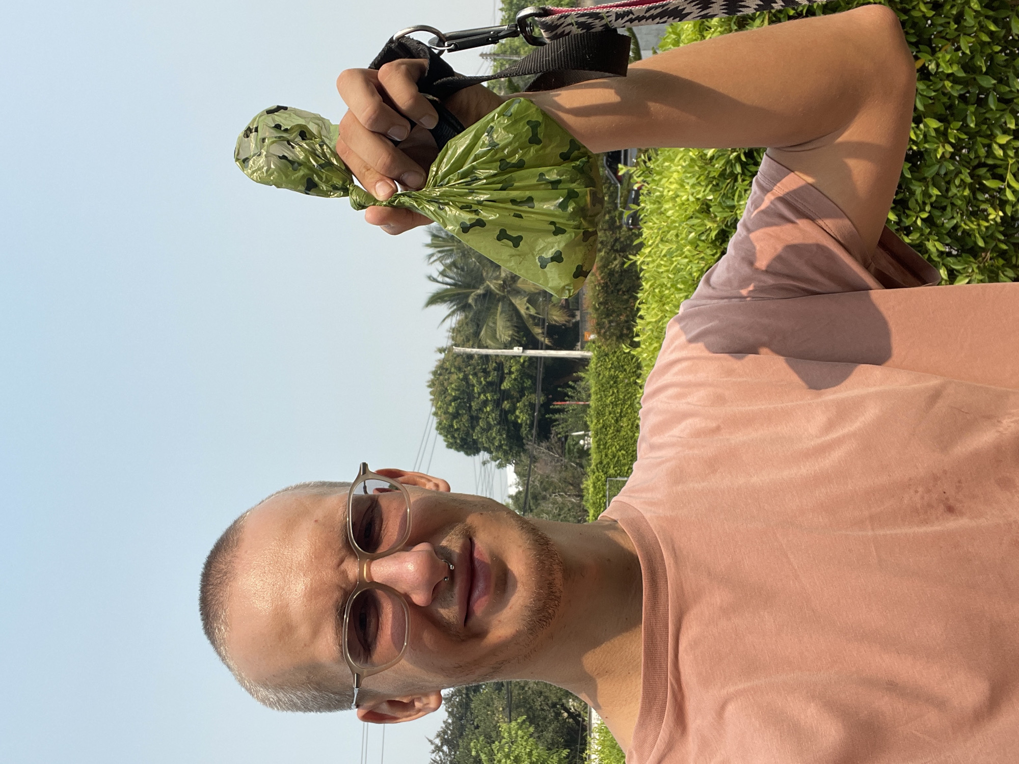 A man with a shaved head and glasses, wearing a pink shirt, holds up a filled green dog waste bag decorated with black bones. He's standing outdoors, possibly in Nong Pa Khrang, Thailand, indicated by the presence of tropical trees and foliage in the background. A dog leash is visible in his hand as well.