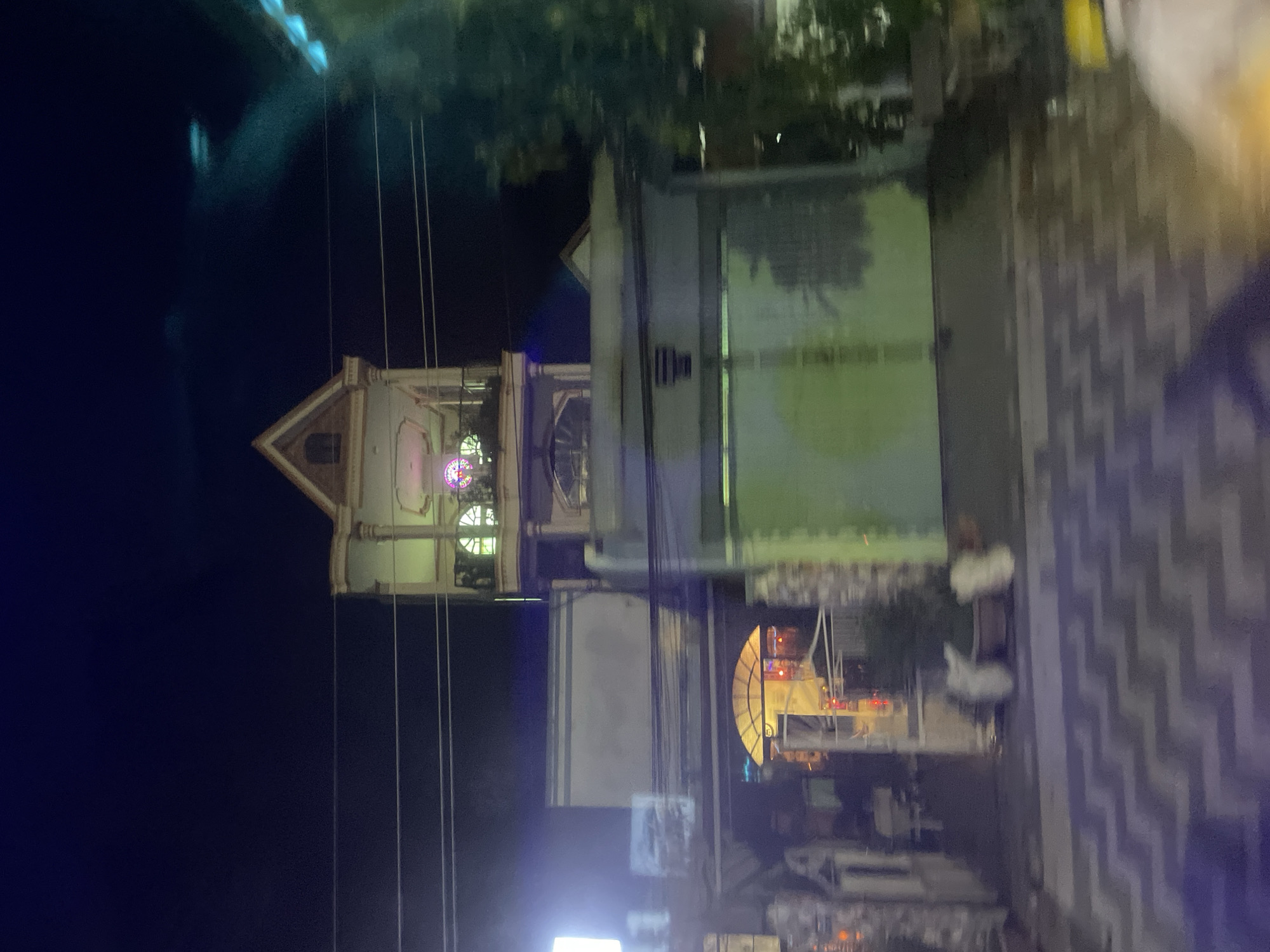 The image captures a dimly lit night scene in Phú Khánh, Vietnam, featuring a multi-story building with a distinct architectural style. The building's facade is a pale color, possibly white or light gray, with decorative elements and arched windows. A prominent feature is a third-story balcony or extension with an ornate window that appears to be illuminated by colorful lights. A triangular roof crowns the uppermost section of the building.
Below this, a large, light-green garage door dominates the ground level of the structure. To the left of the garage door, an entrance is visible, marked by a semicircular archway with yellow lighting. The entrance area shows some clutter, including potted plants and white objects that might be decorative statues or furniture. The ground in front of the building is paved with a zigzag pattern of gray and purple tiles, reflecting the ambient light.
The scene is framed by trees on the right side, partially obscuring the view and adding to the atmosphere of a residential street at night. Power lines are strung horizontally across the upper portion of the image, indicating the presence of urban infrastructure. The overall impression is one of a residential or commercial property in a Vietnamese city after dark, with a mix of traditional and modern architectural influences.