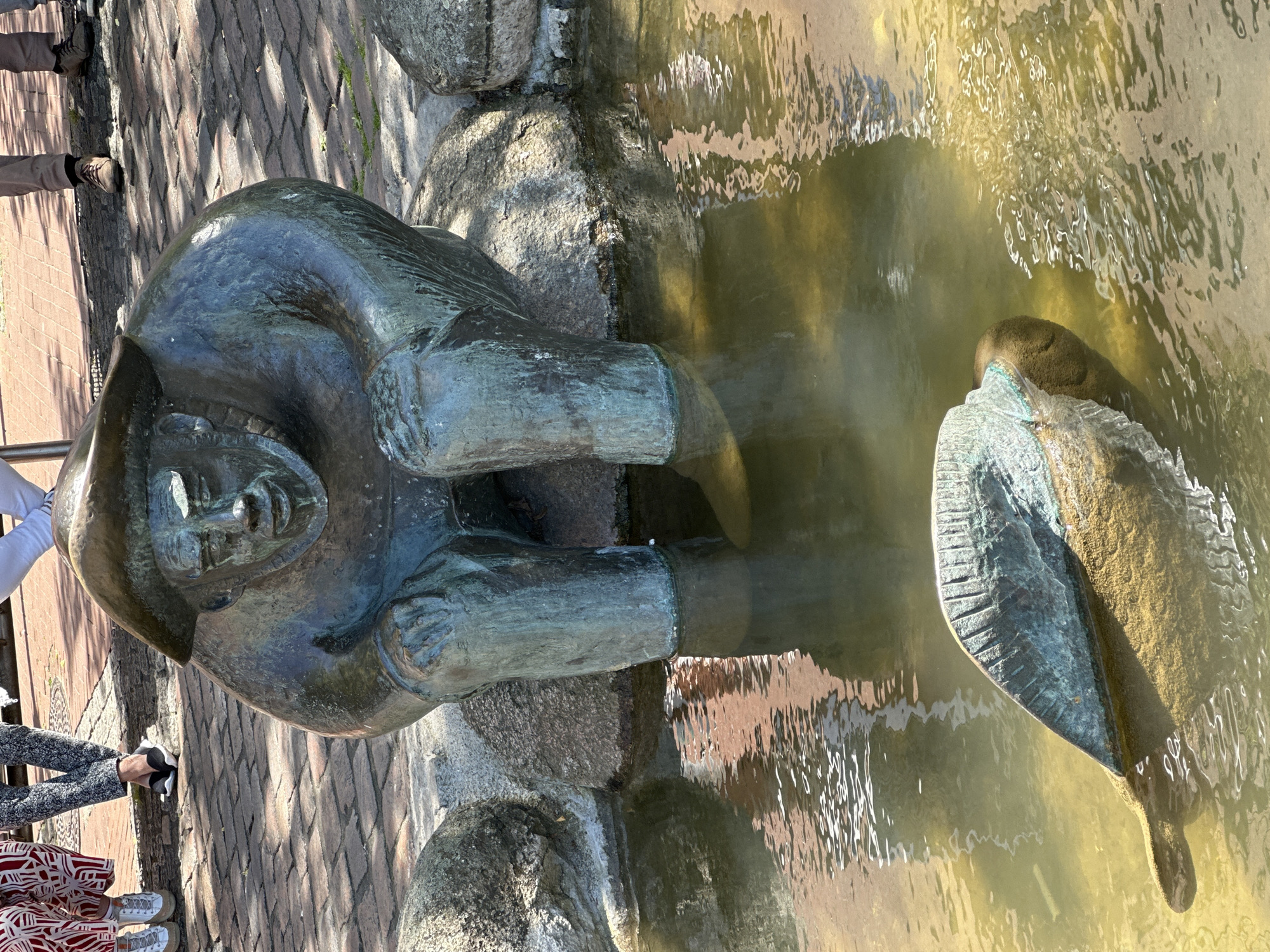 The image depicts a bronze sculpture of a man sitting and dipping his feet in a pool of water in the city of Stade, Germany. The statue is positioned near the edge of a brick-lined area. The water in the pool is murky, with reflections visible. A sculpture of a bird is also in the pool. Portions of at least two people are visible in the periphery.