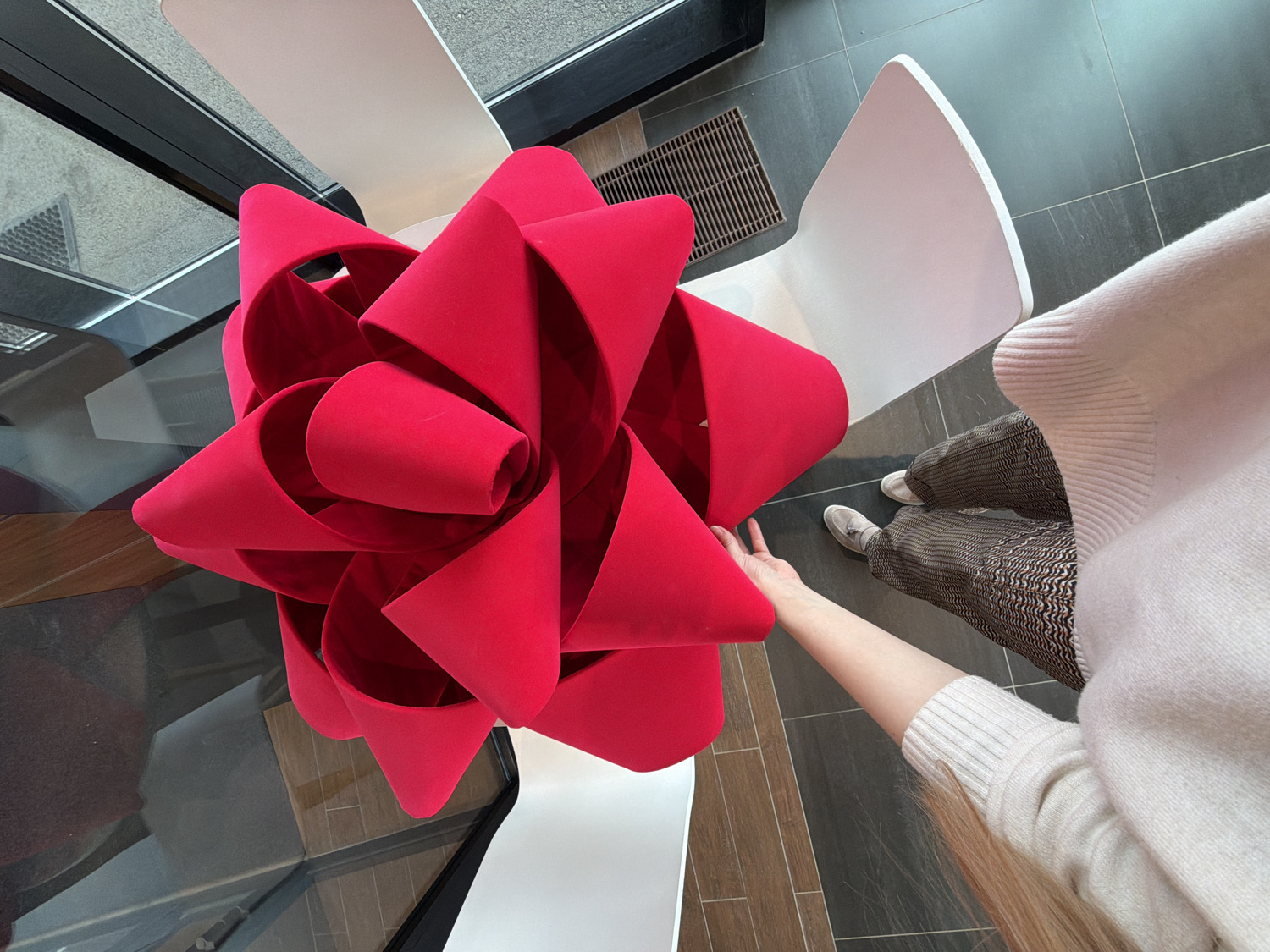The image, taken in Izhevsk, Russia, offers a high-angle perspective of a modern interior space. The primary subject is a large, vibrant red, sculptural object resembling an oversized bow, possibly crafted from a plush material like velvet or felt. Its voluminous loops and folds dominate the center-left of the frame.
A person is partially visible on the right side of the image. Their right arm is extended towards the red bow, with an open hand, suggesting an act of touching or interaction. The person is wearing a light-colored, possibly cream or beige, knitted sweater with ribbed cuffs. Their lower body is clad in brown trousers with a distinctive wavy or zigzag pattern, and light-colored shoes, appearing to be flats or sneakers.
The setting is a contemporary indoor environment, likely a commercial or public lounge area. A dark, reflective surface, possibly a glass table, is present beneath and around the red bow, reflecting parts of the room. Two white, modern chairs with unique, curved designs are visible; one is largely behind the red object, and the other is partially obscured by the person. The floor is composed of dark grey tiles interspersed with sections of wood-patterned flooring. A metal grate, likely an air vent, is visible on the tiled floor. The ambient lighting indicates it is daytime. No visible text is present in the image.