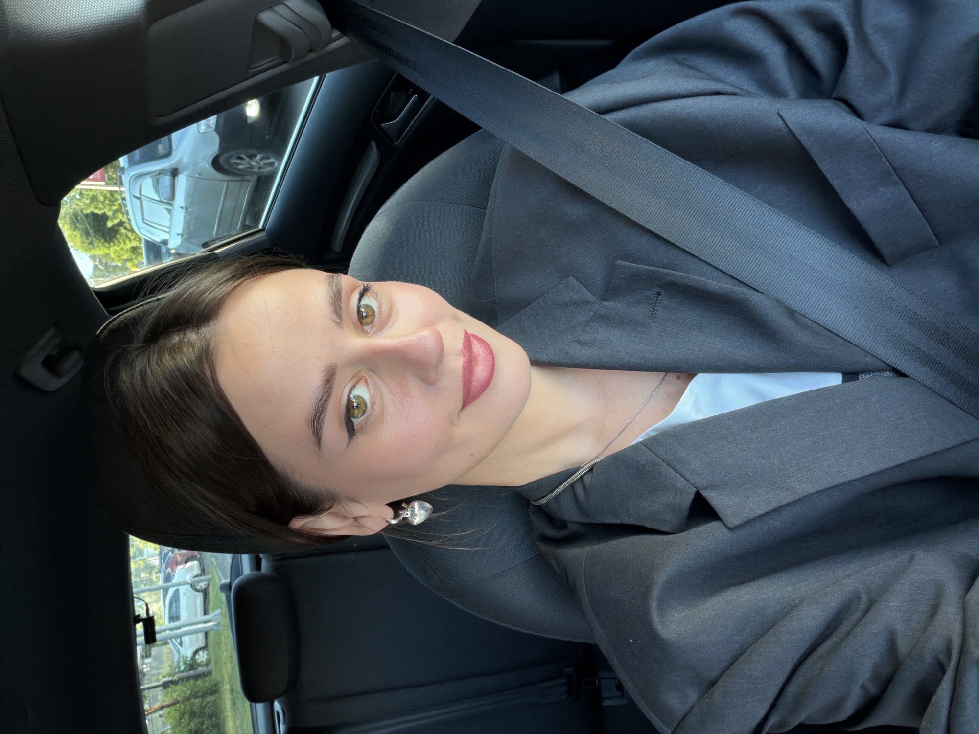 A woman is sitting in the driver's seat of a car, possibly a hatchback, looking at the camera. She has dark hair pulled back and is wearing a black blazer over a white top. Her makeup includes winged eyeliner and pink lipstick. She is wearing a silver heart-shaped earring in her visible ear and a delicate necklace. A seatbelt is visible across her chest and shoulder. The background shows trees and parked cars through the windows, suggesting an outdoor urban environment. The specific location of Krasnoyarsk, Russia, cannot be determined from the visual information in the image.