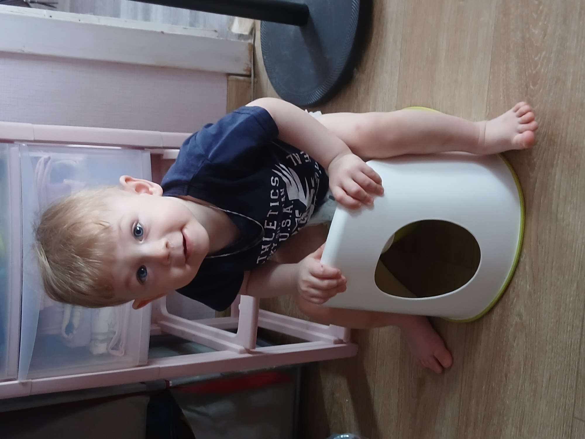 A child, presumably a toddler with blonde hair and blue eyes, is sitting on a white and light green potty. The child is wearing a dark blue shirt with white text. The potty is on a wooden floor. Next to the child is a pink plastic drawer unit. A black metal base of what appears to be a fan is visible behind the child. The scene appears to be indoors. It is impossible to verify the location is in Chaikovsky, Russia from the image alone.