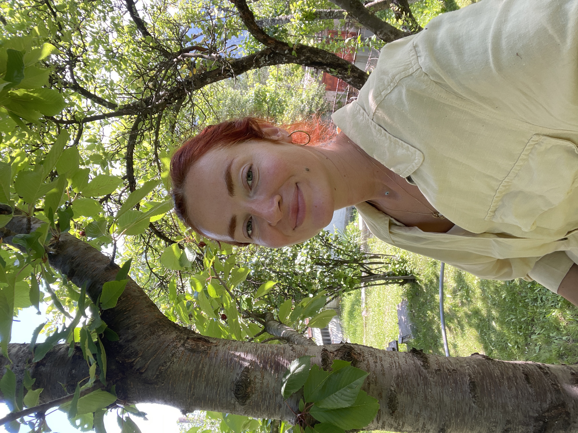 A woman with red hair and fair skin is pictured outdoors, standing next to a tree in what appears to be a garden or yard. She is wearing a light yellow, collared shirt and a necklace. The background shows lush green foliage, with sunlight filtering through the leaves, indicating it is daytime. There are hints of structures and possibly a fence in the distance, suggesting a residential area. The environment is consistent with the description of Rudartsi, Bulgaria, a location known for its natural beauty and greenery.