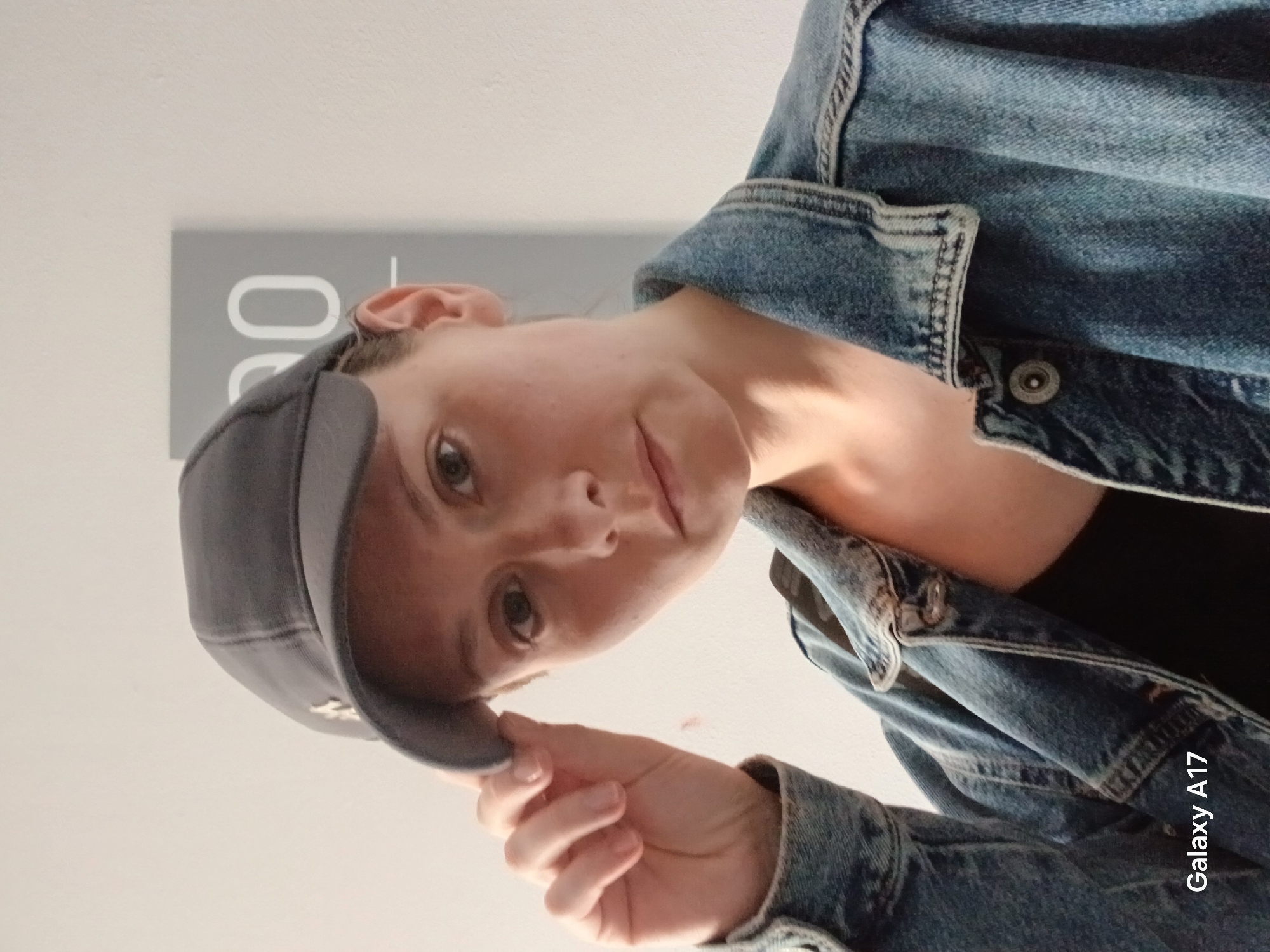 A woman in a denim jacket and gray baseball cap is holding the brim of her cap with her right hand, while looking directly at the camera. She is indoors, with a neutral-toned wall behind her. A gray rectangle with white lettering is partially visible in the upper left corner. The image was taken with a Galaxy A17. No location is available.