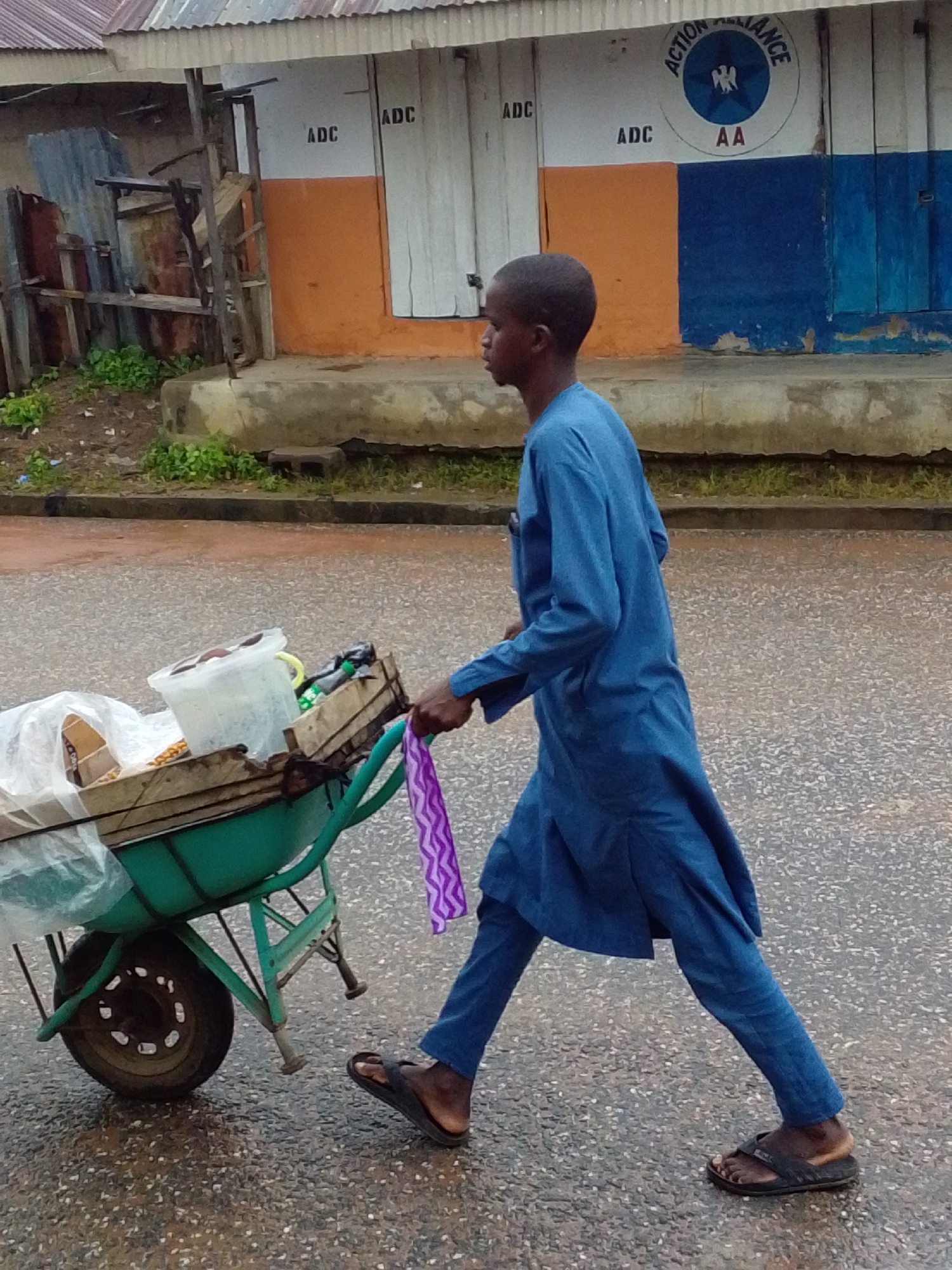 A young boy in a blue tunic and pants walks to the right, pushing a green wheelbarrow filled with items, including a white bucket and a plastic bag, along a wet asphalt road. In the background, a building with a corrugated metal roof has the words "ADC" and "ACTION ALLIANCE AA" painted on its walls. The ground is wet, suggesting recent rain.