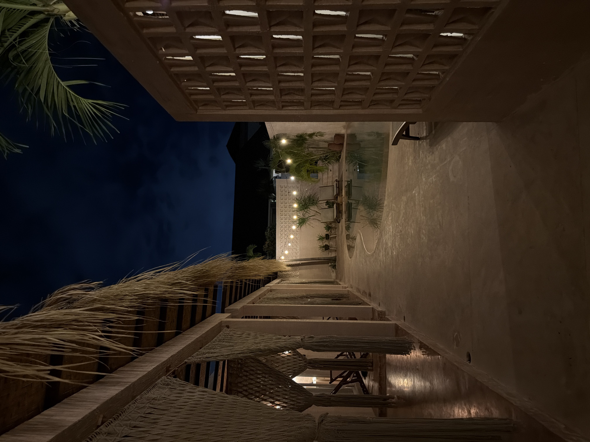 This nighttime image appears to be taken in a restaurant or bar in North Kuta, Indonesia. The location features open-air architecture with visible concrete lattice roofing and thatched-roof elements. String lights illuminate a central area with seating and possibly a water feature (pool or decorative basin) visible in the distance. The night sky is dark. There are no discernible people in the shot.