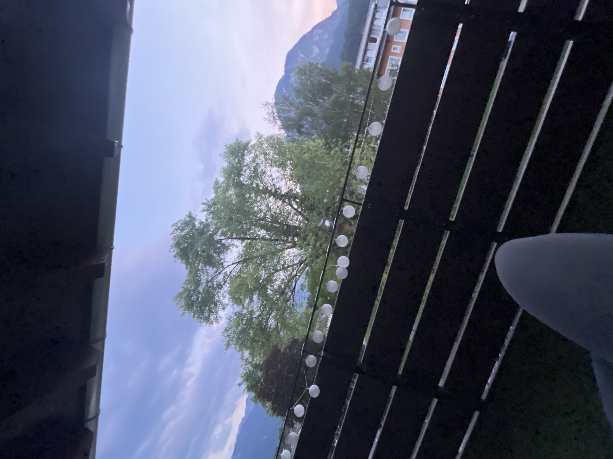 The image depicts an outdoor scene viewed from a balcony or patio, looking out towards mountains and trees. A dark wooden railing with horizontal slats runs along the right side of the frame, adorned with a string of white globe lights. The sky is visible in the background, with clouds and what appears to be the soft hues of either sunrise or sunset, suggesting early morning or late evening.
Beyond the railing, lush green trees dominate the midground, with a larger, mature tree in the center of the frame. In the distance, a majestic mountain range is visible, its peaks softened by the atmospheric light. To the right of the mountains and trees, a building with multiple windows and an orange facade can be seen, indicating a residential or commercial area. The overall impression is one of a peaceful, scenic view from a comfortable vantage point, possibly a home in a mountainous region.
The perspective is from a low angle, looking upwards, with a dark overhang on the left side suggesting the edge of a roof or balcony above. The lighting is diffused, consistent with twilight conditions. There are no discernible people or specific activities taking place, suggesting a still moment in time. The presence of the string lights implies the space is set up for evening relaxation. No text is visible in the image. The location context of Garmisch-Partenkirchen, Germany, aligns with the mountainous landscape and architecture.