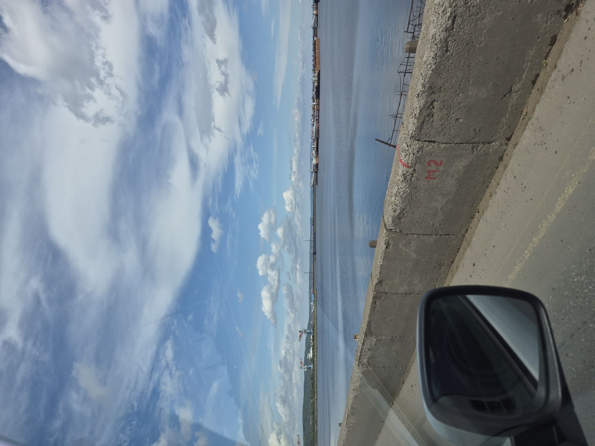 This image shows a wide expanse of water, likely a river or reservoir, with a distant shoreline featuring industrial elements. In the foreground, a concrete barrier runs along a road, with a car's side mirror visible on the left. The sky is blue with scattered clouds, suggesting a daytime scene.
The location is the Kama River in Chaikovsky, Russia. The industrial structures on the distant shore, including cranes and what appear to be port facilities or factories, are characteristic of the city's industrial landscape. The presence of numerous ships or barges in the water indicates ongoing maritime activity. The concrete barrier and road suggest this is a view from a bridge or a road alongside the water. The number "M2" is spray-painted in red on the concrete barrier.