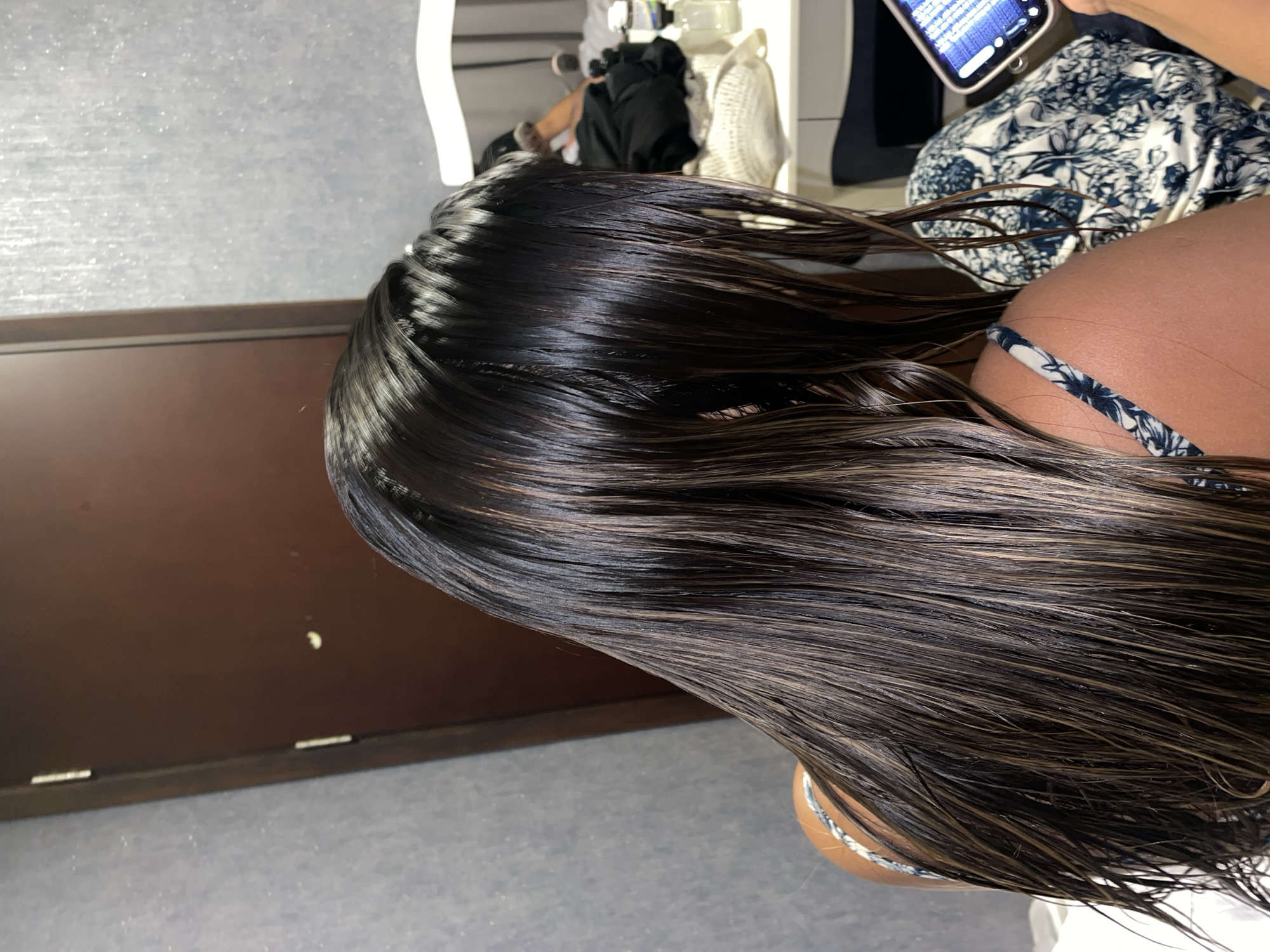 A close-up, slightly tilted, medium shot captures a person with wet, dark hair, featuring lighter brown highlights. The person's shoulder, clad in a blue and white floral-patterned bikini strap, is partially visible. A mobile phone is seen near the subject's head, possibly being used to take the picture. Background elements include a dark brown cabinet or surface adjacent to a light grey floor. The setting appears to be indoors, perhaps in a bathroom or changing room. The image does not provide information on which to place the image in Central Jakarta, Indonesia.