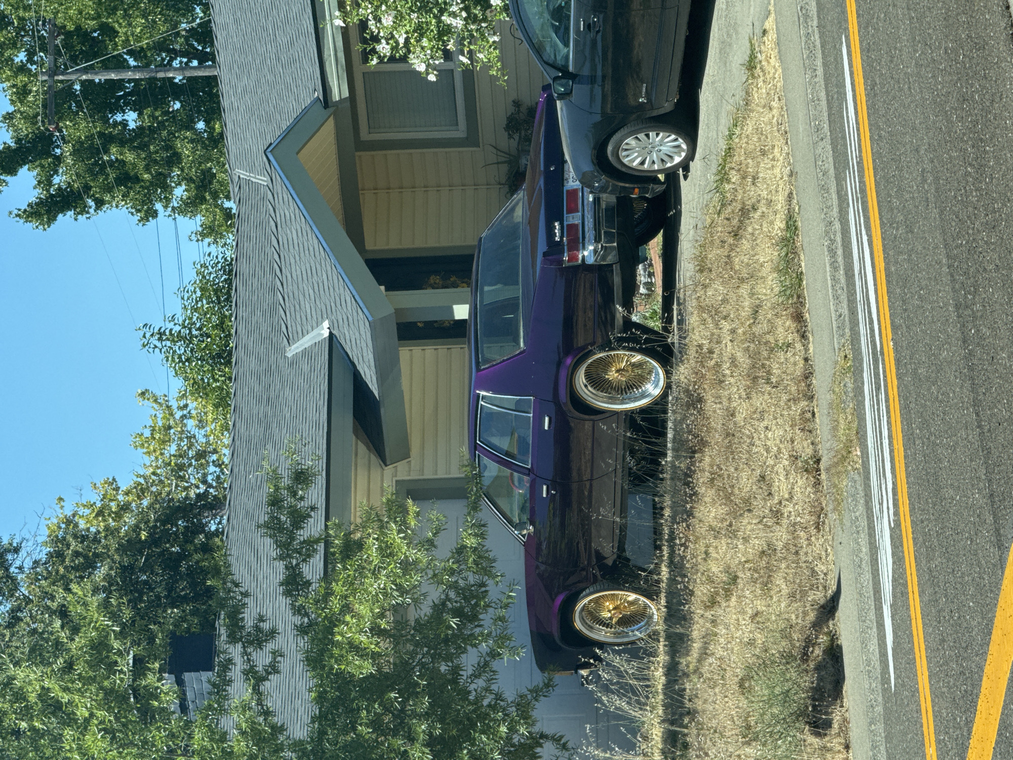 An eye-level, medium shot captures a vintage purple sedan with gold wire wheels parked on the side of a residential street in Citrus Heights, USA. The car is positioned in front of a beige, single-story house with a gray shingled roof. To the right of the purple sedan, a dark-colored hybrid vehicle is partially visible, also parked on the street. The foreground shows a curb and a paved road with yellow and white lane markings. Dry, yellowed grass and some green bushes cover the area between the curb and the house. Lush green trees and a clear blue sky form the background.