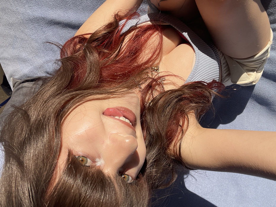 An individual is depicted in a high-angle, close-up perspective, lying on their back on a light blue textured surface. The subject has long, dark brown hair with reddish-auburn streaks, particularly visible on the viewer's right side of their head. Their light brown/hazel eyes are open, and their mouth is open with the tongue slightly protruding from the left side. Strong, direct sunlight illuminates the subject, creating specular highlights on the skin (forehead, nose) and casting distinct shadows onto the blue surface. The individual wears a light blue ribbed camisole, revealing a prominent cleavage line, and partially visible light-colored shorts or an undergarment. A silver-toned necklace adorned with multiple small charms is visible around the neck. A dark, intricate tattoo is partially visible on the subject's left upper arm/shoulder area. The scene is located in the city of Raanana, Israel.