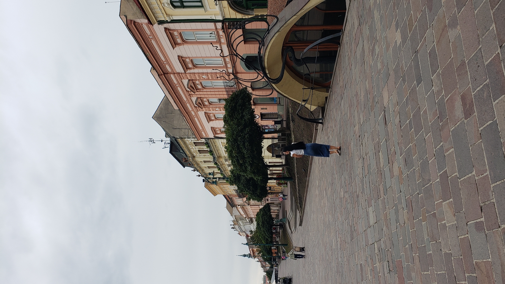 The image depicts a street scene in Prešov, Slovakia, specifically the pedestrian zone of Hlavná Street. The perspective is looking down the street from a slightly elevated viewpoint. The street is paved with cobblestones and lined with historic buildings with colorful facades, featuring ornate architectural details. Several trees are planted along the sidewalk, providing shade. In the foreground, a woman with dark hair is standing on the cobblestone street, wearing a white top and a long blue skirt. She is looking away from the camera. Further down the street, a person is pushing a wheeled cart, and other individuals can be seen walking. The sky is overcast, suggesting a cloudy day. The architecture of the buildings, with their multi-story designs and decorative elements, indicates a historical European city center. The unique, curved entrance on the right side of the frame, with its metal artistic embellishments, is a distinctive feature of the scene.