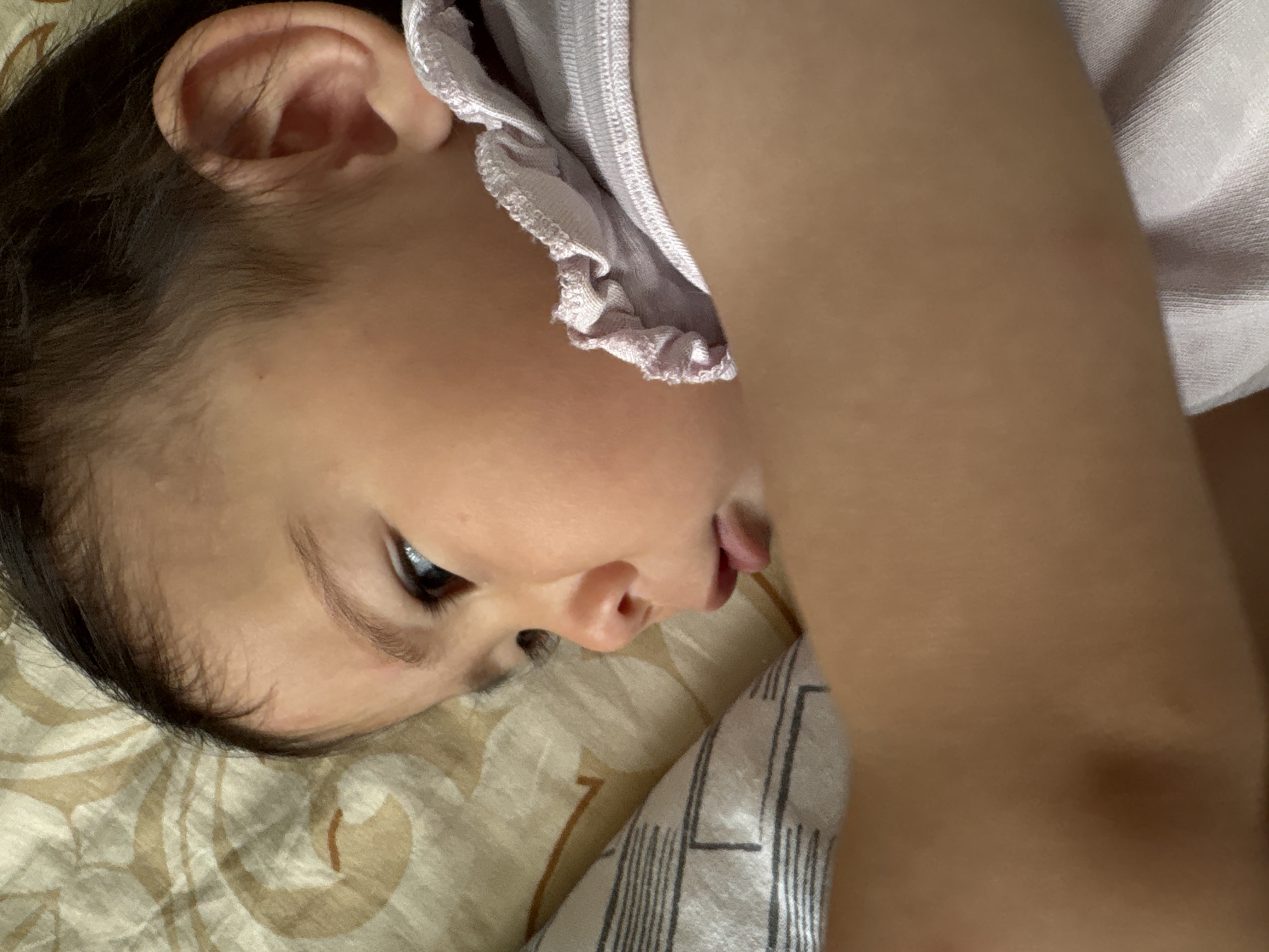 A young child with dark hair lies on a bed with patterned bedding, possibly in Ембаево, Russia. The child is wearing a light-colored shirt with a ruffled neckline. The child's face is turned to the side, with one eye visible. A portion of the child's arm is also visible.