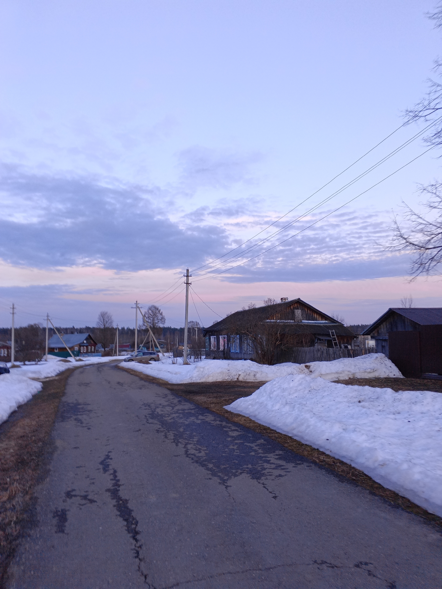 The image shows a rural scene in Красный Маяк, Russia. An asphalt road leads into the distance, with patches and cracks visible. Patches of melting snow are present along the road's edges and in front of houses. Houses are scattered along the road, with traditional wooden architecture. Power lines stretch across the sky above the road. The sky is overcast with a mix of blues, grays, and hints of pink, suggesting either dawn or dusk. Bare trees are visible on the right side of the frame.