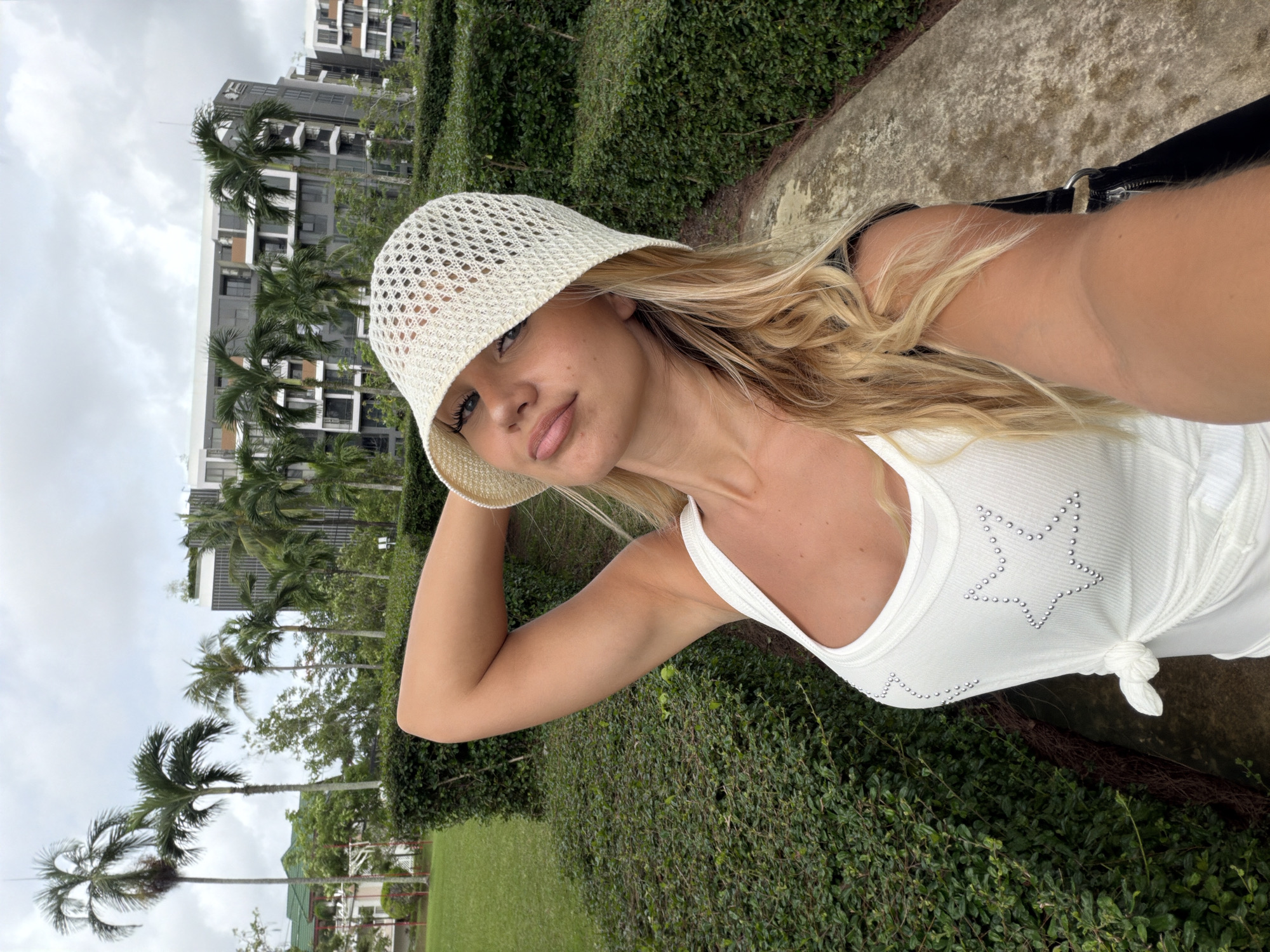 A young Caucasian woman with blonde wavy hair and blue eyes is taking a selfie outdoors in a garden setting. She is wearing a white knitted bucket hat and a white tank top adorned with silver studs in the shape of stars. Her left arm is raised behind her head, and her right arm is holding the phone to take the picture. She is wearing a dark-colored bag over her right shoulder. The background features neatly trimmed green hedges, a stone pathway, lush green grass, and several palm trees. In the distance, modern apartment buildings with gray facades and balconies are visible. The sky is overcast with clouds. The location is the city of Choeng Thale, Thailand.
