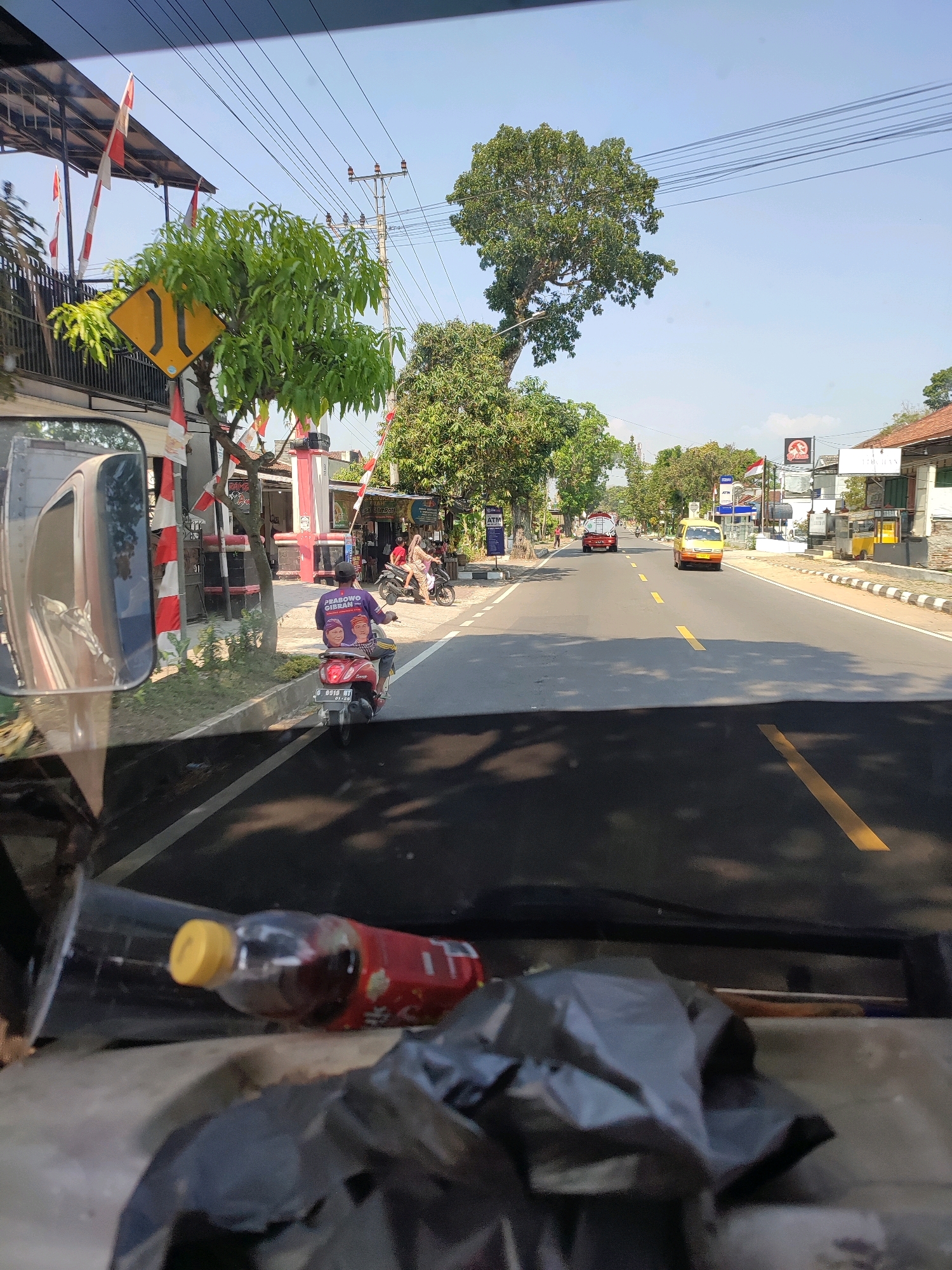 The image shows a street scene in Kemendangun, Indonesia, on a sunny day. The perspective is from inside a vehicle, looking forward through the windshield and side mirror. The road is paved with a white and yellow dashed line in the center. On the left side of the road, there is a sidewalk with trees, a building with red and white striped flags, and a yellow diamond-shaped road sign indicating a narrowing of the road. A person is riding a red scooter wearing a purple shirt with a picture of faces on it. A red bottle with a yellow cap lies on the dashboard in the foreground. On the right side of the road, there are buildings, a yellow taxi, and a white truck. The sky is clear and blue.