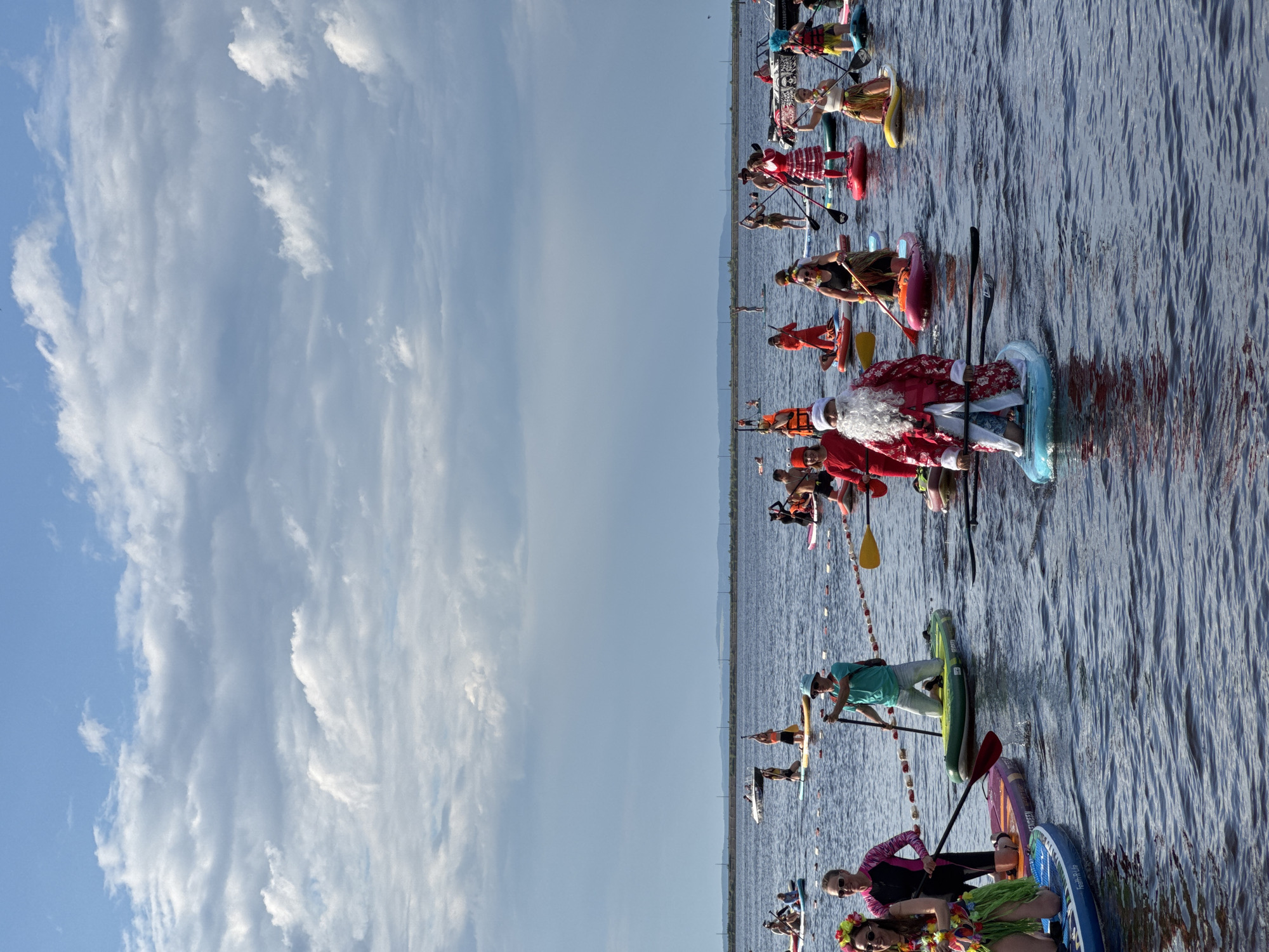 The image shows a group of people on paddleboards in a body of water. Many of the participants are dressed in festive attire, including a person dressed as Santa Claus, people in Hawaiian-themed outfits with leis and grass skirts, and others in red and white outfits. Some are wearing life vests. The people are spread out across the water, some paddling and others appearing to be stationary.
The setting is an outdoor scene with a clear sky filled with wispy white clouds on the left side of the image. The body of water appears to be a lake or a wide river, with a distant shoreline visible. The water is rippled, suggesting a gentle breeze. The light in the image suggests it is daytime, likely late afternoon or early evening given the soft lighting.
The event appears to be a festive paddleboarding gathering or parade, possibly related to a holiday celebration. The mood seems to be joyful and celebratory, with participants embracing the festive costumes and participating in a shared activity. The presence of colorful paddleboards and decorations (like the string of small flags or pennants) adds to the celebratory atmosphere. No visible text is present in the image.