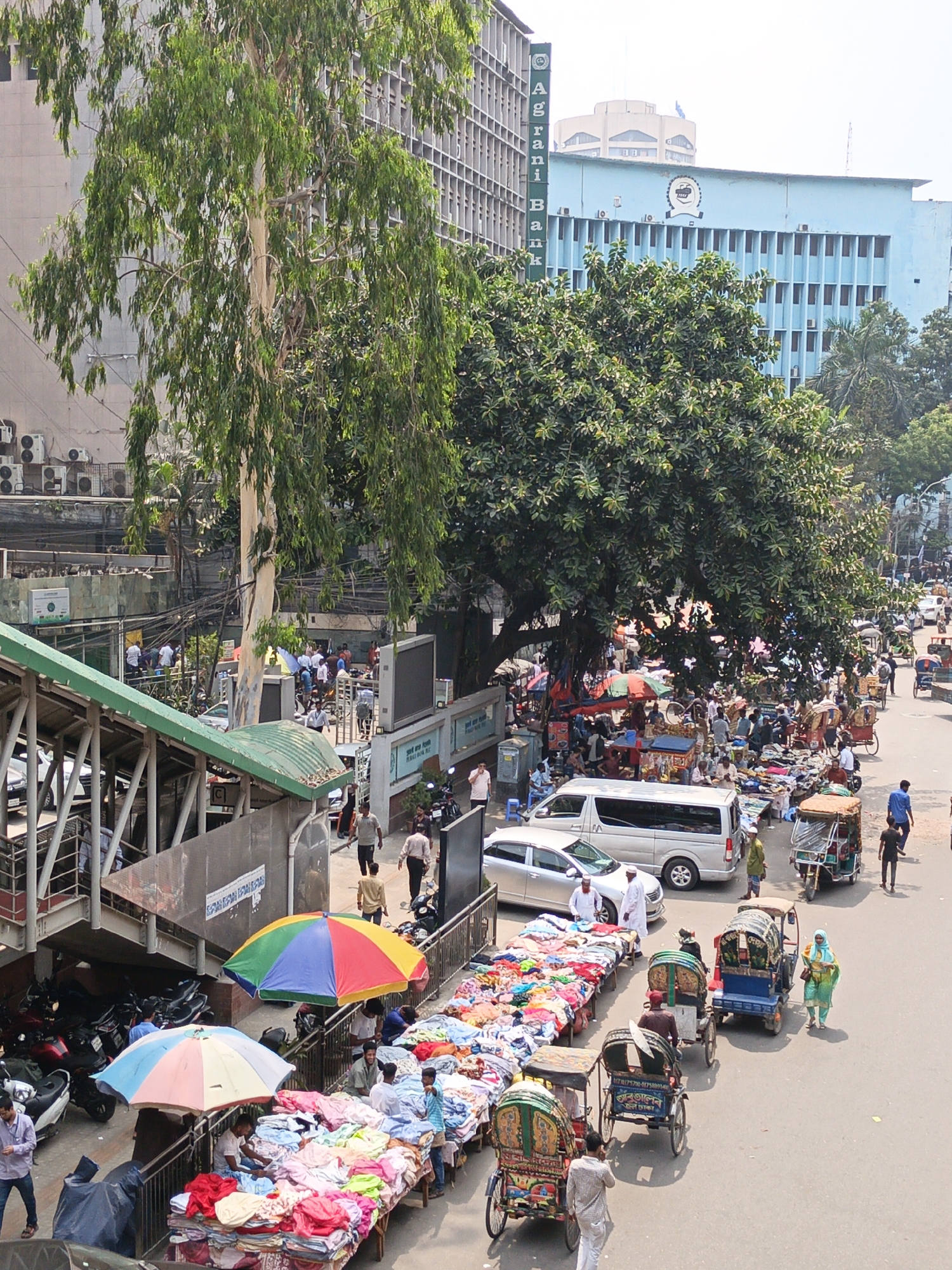 This is a street scene in Dhaka, Bangladesh. A street market, with vendors selling colorful textiles, is set up on the road. People are shopping and interacting with the vendors. Rickshaws and auto-rickshaws navigate the crowded street. Pedestrians walk along the sidewalks and cross the road. In the background are large buildings, including one that says "Agrani Bank" on the side. There is a footbridge on the left with motorcycles parked nearby. The scene is active and bustling with people and traffic.