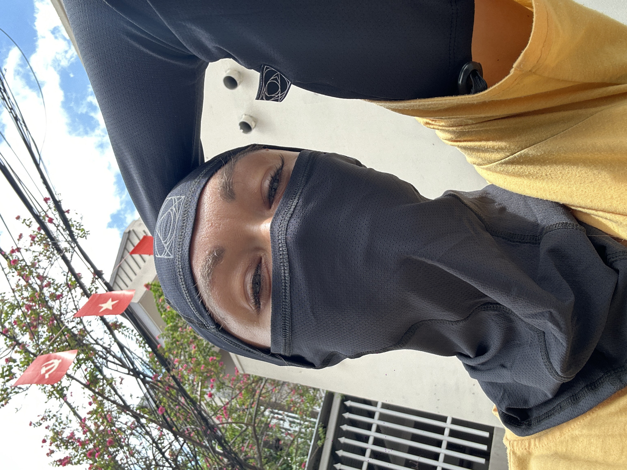 A person wearing a black balaclava-style head covering that obscures the lower half of the face, revealing only the eyes, is positioned outdoors. Above the person, a black long-sleeved shirt with a tag near the shoulder is visible. To the right, a yellow garment is present. The background features red flags with white stars or symbols, possibly political or nationalistic emblems. Lush green trees with pink blossoms and power lines are also visible. Architectural elements, like a white wall and a metal grate, are present. It appears to be in the city of Đồng Nai, Vietnam, indicated by the flags.