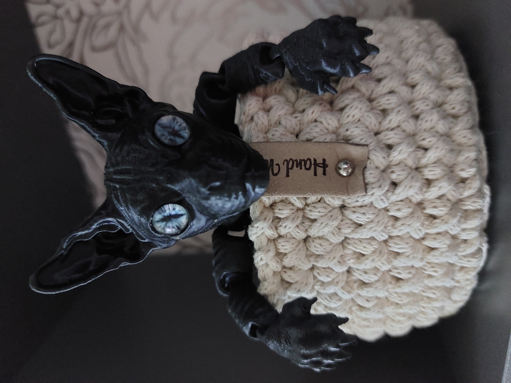 A close-up shot depicts a handcrafted doll resembling a sheep with a black, skeletal-like creature emerging from its center. The sheep's body is constructed from tightly crocheted yarn in a light, off-white color. Affixed to its side is a rectangular tag with the handwritten word "Hand" visible, along with two metal rivets. The head and limbs of the creature are black, with pronounced joints, long ears, and detailed claws. Its eyes are a striking light blue with star-like patterns. The creature's head is prominently positioned to the left, appearing to emerge from the sheep's body. The background consists of a dark surface and a blurred, patterned fabric. There are no indicators that this photo was taken in Chaikovsky, Russia.