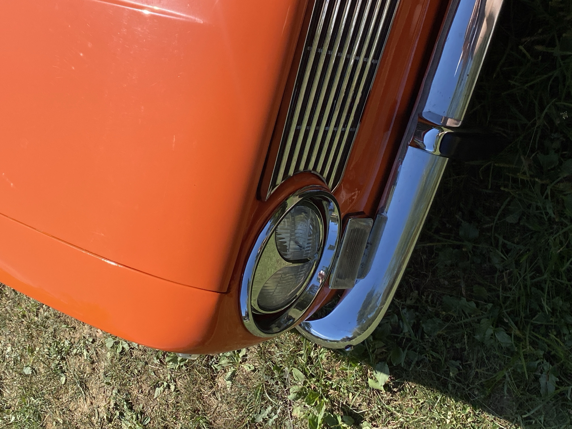 The image shows a close-up, eye-level view of the front right side of a vintage orange car, likely parked on grass in Bezymyannoe, Russia. The car's hood, headlight, grille, and chrome bumper are the primary subjects. The grass is green and dry in some patches, with scattered brown leaves and weeds, suggesting an outdoor setting, possibly a yard or park. The lighting indicates it is daytime, with a shadow cast across the grass from the car. No people are visible in the frame.