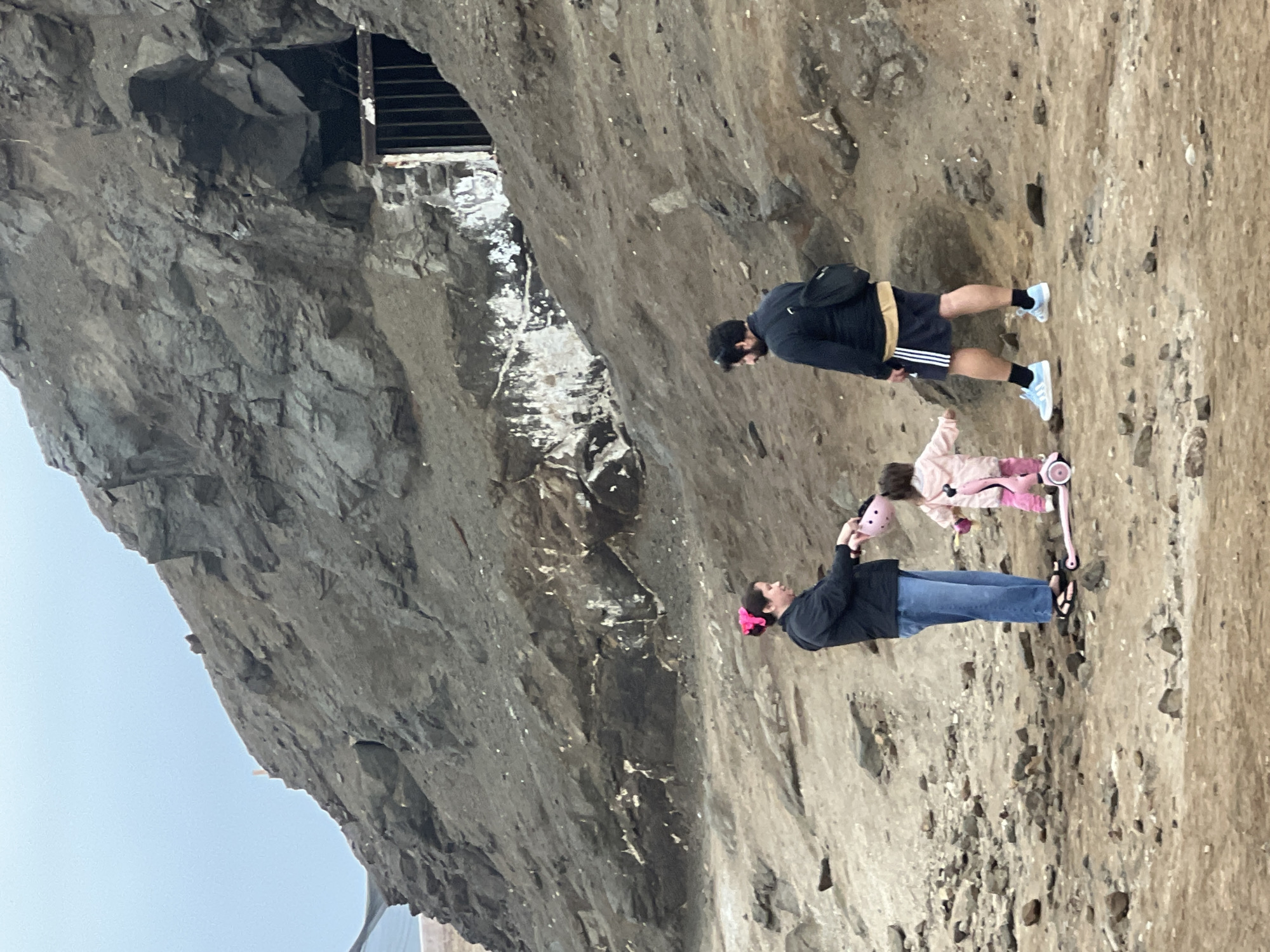 In the city of Arica, Chile, a family stands near the entrance of a cave at the base of a rocky cliff. A woman, wearing a black jacket and blue jeans, holds a pink helmet. A young child, dressed in a pink jacket and pants, rides a pink scooter. A man, wearing a black jacket, shorts, and athletic shoes, stands beside them, with a backpack on his back. The cave entrance is secured with a metal grate. The ground is covered in rocks and dirt, and the sky is overcast.