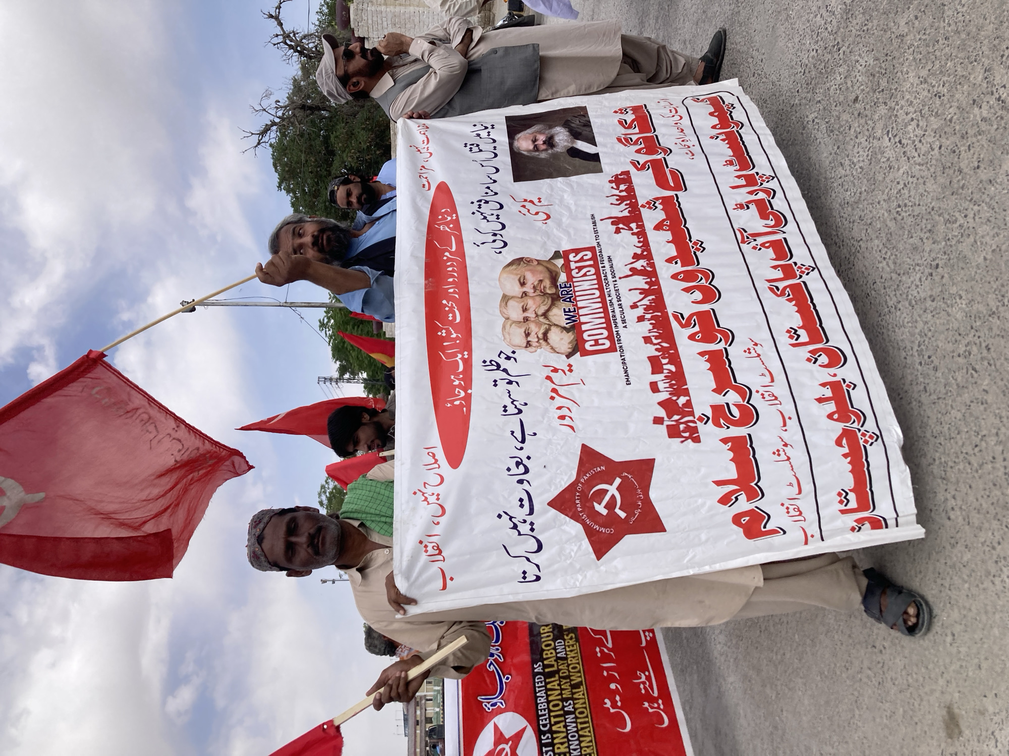 The image shows a street scene in Quetta City Tehsil, Pakistan, featuring a group of people holding banners and flags in what appears to be a political demonstration. The banners are predominantly white with red lettering in Urdu, and English text. One banner has portraits of Karl Marx and three other men, the hammer and sickle symbol, and the text "WE ARE COMMUNISTS." Another banner proclaims "INTERNATIONAL LABOUR" and mentions "MAY DAY". The individuals include men wearing traditional Pakistani clothing. Some are holding red flags, also adorned with the hammer and sickle symbol. The sky is visible with scattered clouds. The setting appears to be urban, with a paved road and buildings in the background.