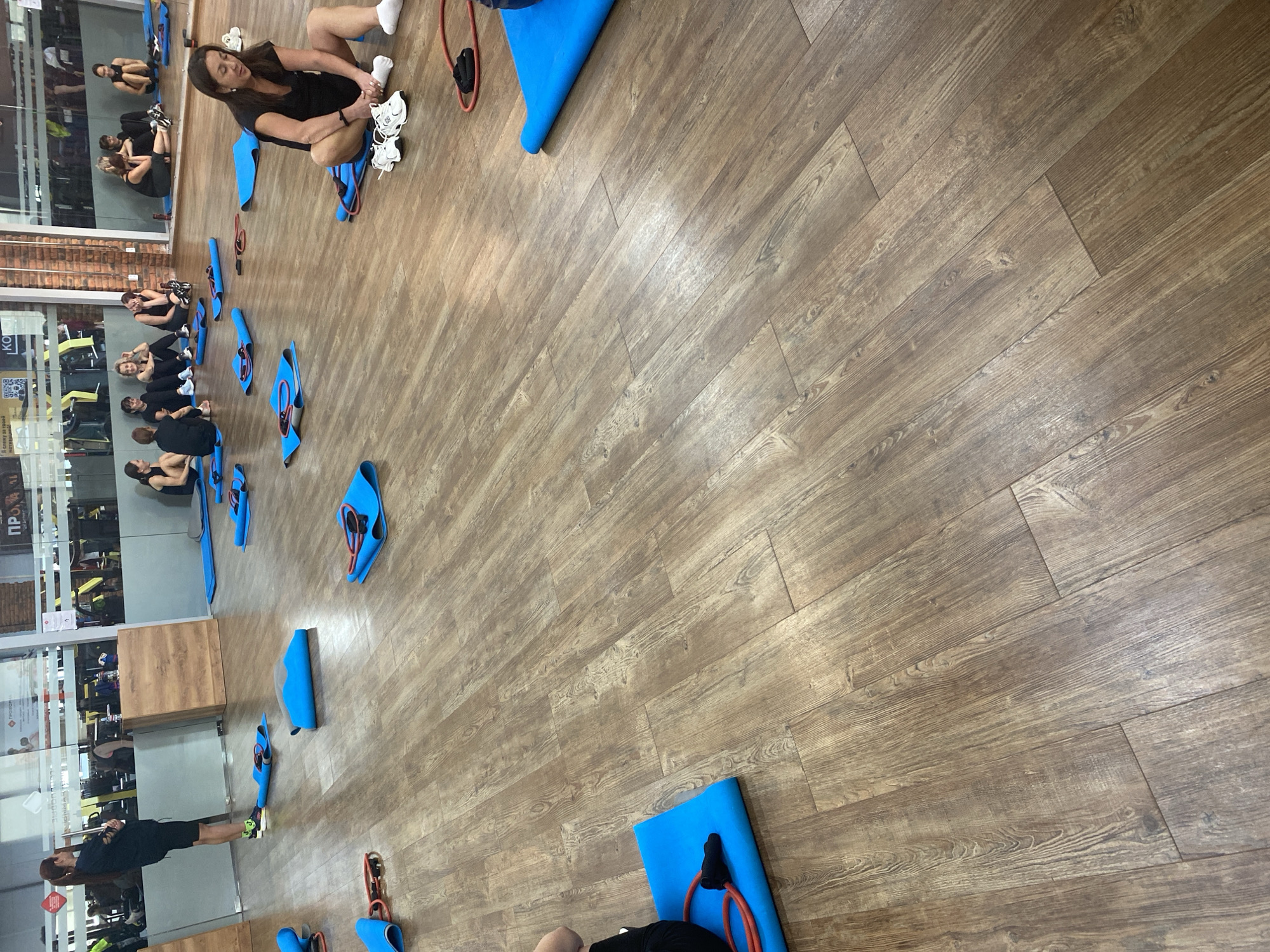 The image depicts an indoor gym with a wooden floor. Several blue exercise mats are scattered across the floor, some with red resistance bands. In the foreground, a woman is sitting on a blue mat, wearing black athletic clothing and white sneakers. Another woman is standing in the background, also wearing black athletic attire. Several other women are visible sitting on mats in the middle ground and background, all dressed in athletic wear. The gym environment includes large windows with reflections of gym equipment and mirrors. The lighting suggests an indoor setting.