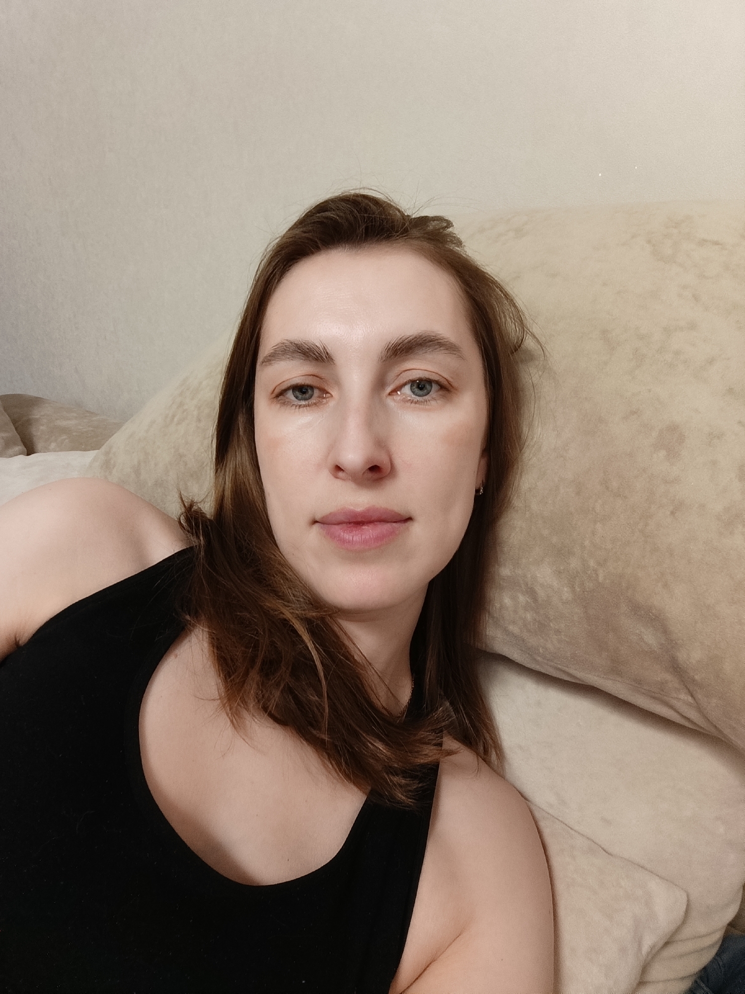 A young woman is lying down, presumably on a bed or sofa, as indicated by the beige cushion behind her head. She is wearing a black tank top. The woman has light skin, light brown hair, and light blue eyes. Her expression appears relaxed. The background is a plain beige wall. The image location, Chaikovsky, Russia, is not visually apparent in the image. There is no evidence within the image to confirm that location.
