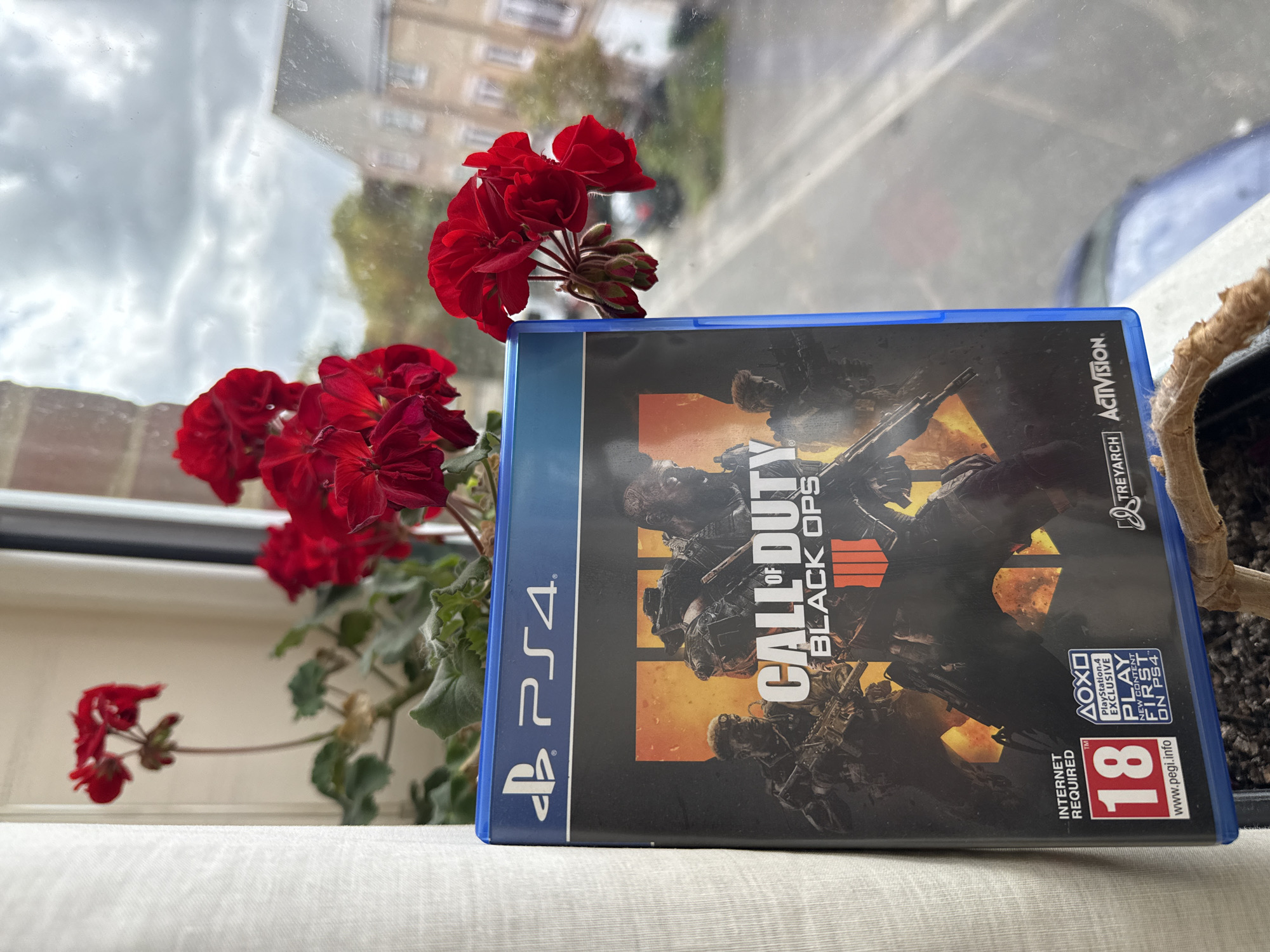 A PlayStation 4 game case for Call of Duty: Black Ops 4 is resting on a white surface, likely a windowsill, next to a potted red geranium. The game case is predominantly black with orange accents and features the game's title prominently. A blue PS4 logo is visible on the left side. The game rating logo is also visible. The geranium has vibrant red flowers. Through the window behind the game case and flowers, a cityscape can be seen under a cloudy sky. The location is within the city of Great Yarmouth, United Kingdom.