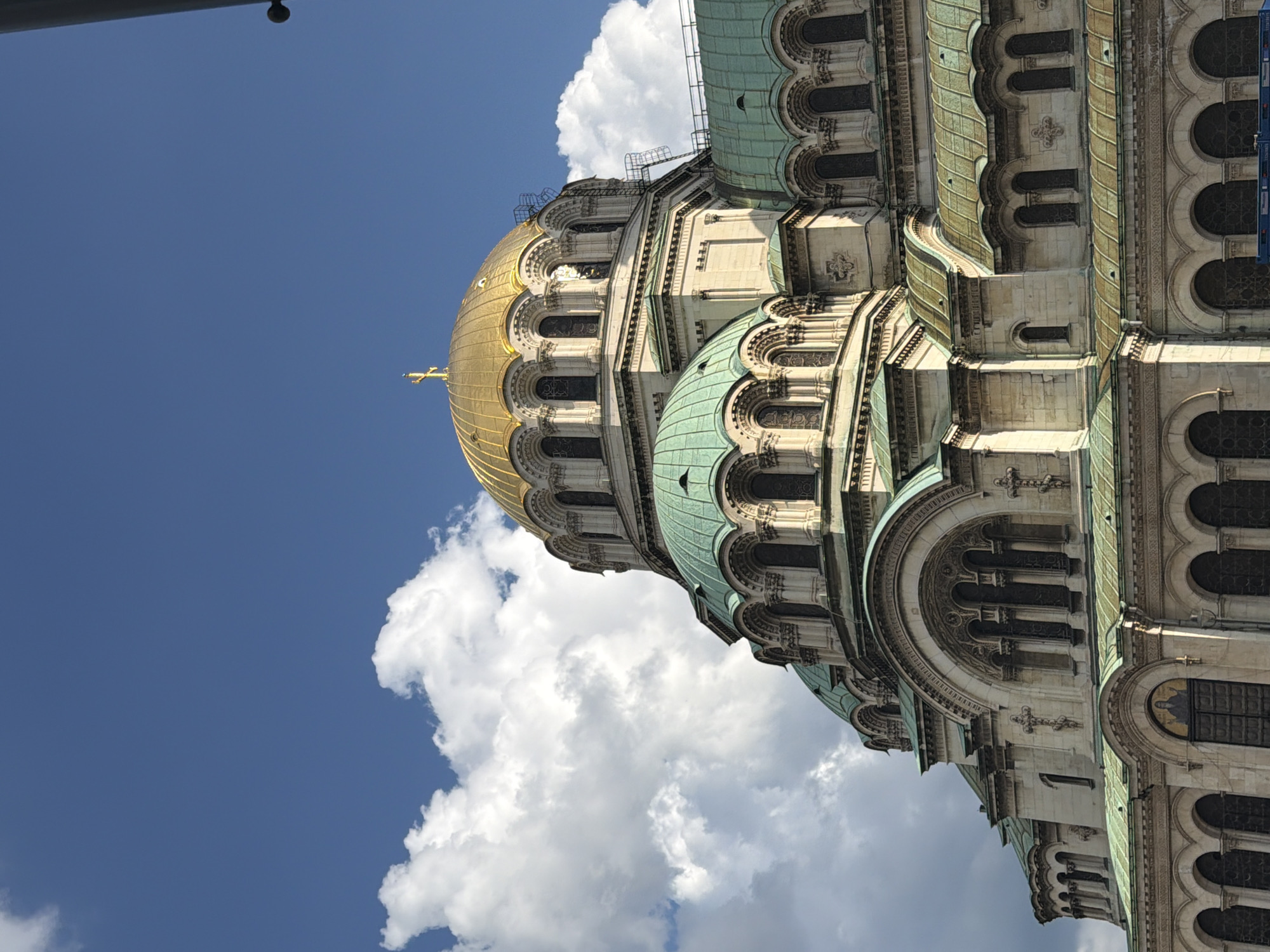The image presents a detailed, upward-angled view of the Alexander Nevsky Cathedral in Sofia, Bulgaria. The central feature is a large, golden dome, crowned with an Orthodox cross, positioned against a clear blue sky with scattered white cumulus clouds. Below and around this main dome, multiple smaller domes and roofs are visible, all covered in verdigris-patinated copper, displaying a distinctive teal-green color. The cathedral's facade is constructed from light-toned stone, featuring intricate architectural details, including rows of arched windows, decorative carvings, and sculpted crosses. Scaffolding is observable on the right side, extending up towards the central golden dome. A religious mosaic or fresco is discernible above an arched entrance at the lower visible section of the building. The scene is illuminated by bright natural light, creating subtle shadows that define the architectural elements. The very bottom right corner of the frame reveals a small, indistinct blue object. No individuals, animals, or specific actions are depicted within the image.