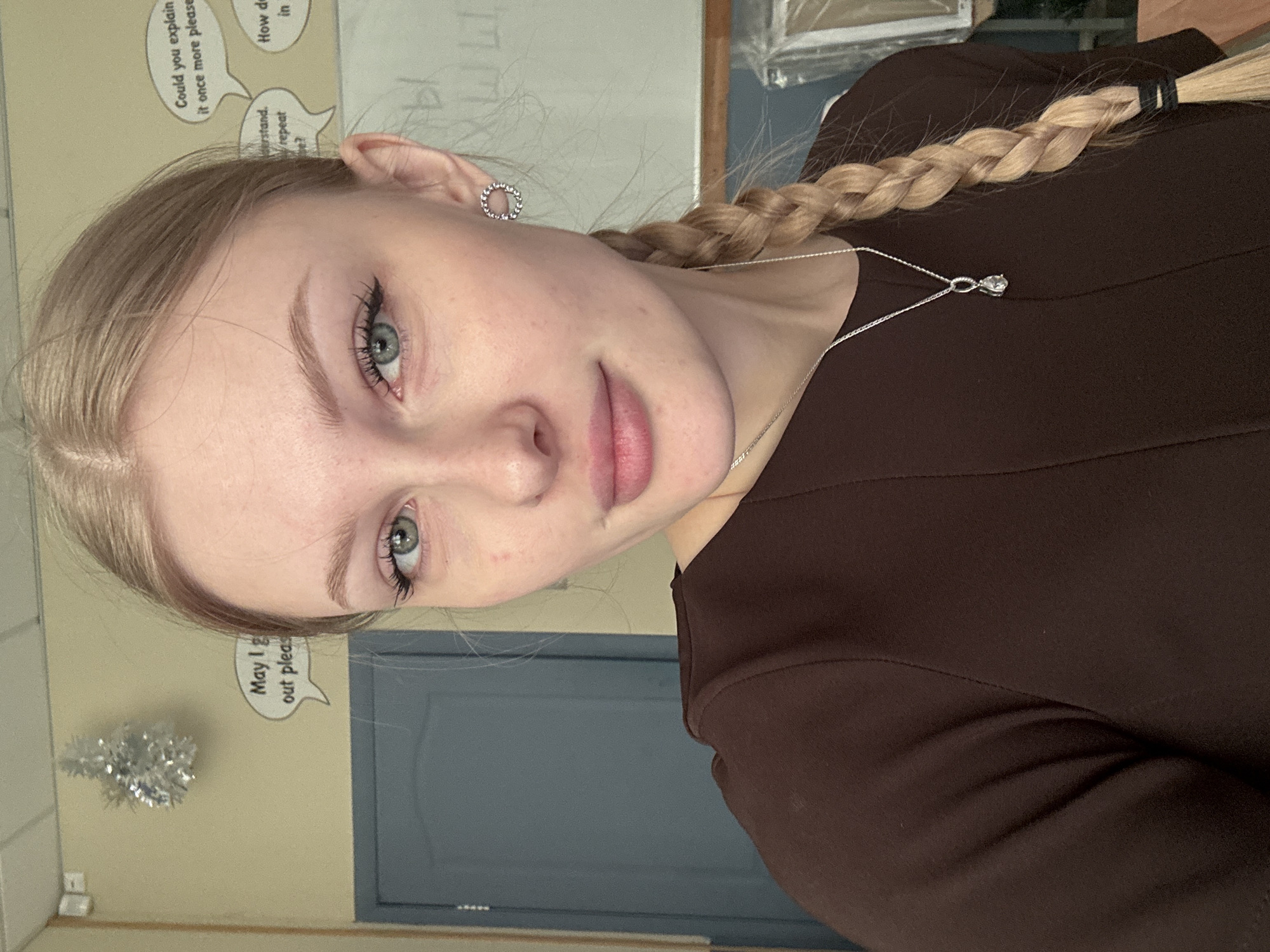 A young, fair-skinned woman with blonde hair braided on the right side of her face and adorned with a small silver necklace poses for a selfie. She is wearing a dark brown dress. Her left ear is visible, featuring a round silver earring. The background includes a tan wall with educational speech bubble stickers ("Could you explain it once more please", "Understand, repeat?", "How do in"), and a silver tinsel decoration. A door or wall section is visible in the background, painted a muted blue-grey. There is no information in the image or the OCR to suggest the location is in the city of Perm, Russia.