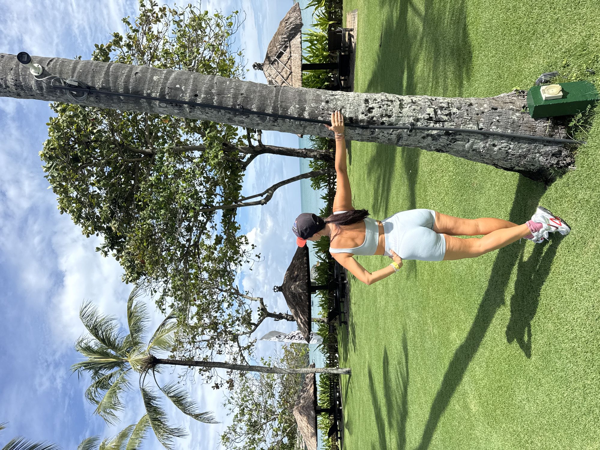 The image captures a woman performing an outdoor fitness pose against a large tree trunk in a tropical setting. She is viewed from behind, wearing a light blue sports bra, matching athletic shorts, a dark baseball cap, and white sneakers with pink accents (the New Balance logo is visible on her left shoe). Her right arm is extended against the tree trunk, and her body is in a plank-like position, with her legs stretched back parallel to the ground.
The setting features a meticulously maintained green lawn, possibly artificial turf, under a bright blue sky dotted with scattered white clouds. The backdrop includes a body of water, likely the ocean or a bay, indicating a coastal location. Several lush green trees, including tall palm trees, are spread across the landscape. In the mid-ground, traditional thatched-roof huts or gazebos are visible, along with outdoor furniture.
Notable details include a white security camera mounted high on the tree trunk the woman is leaning on, with a black cable running down. At the base of the tree, a dark green box with a small circular white object on top is present. Long shadows from the trees and the woman are cast across the grass, suggesting it is a sunny day, likely late morning or afternoon. A partially visible white flag or banner in the background shows the text "THE BE". The overall scene evokes a resort-like atmosphere in a beautiful, sunny tropical location, consistent with Kuta Selatan, Indonesia.