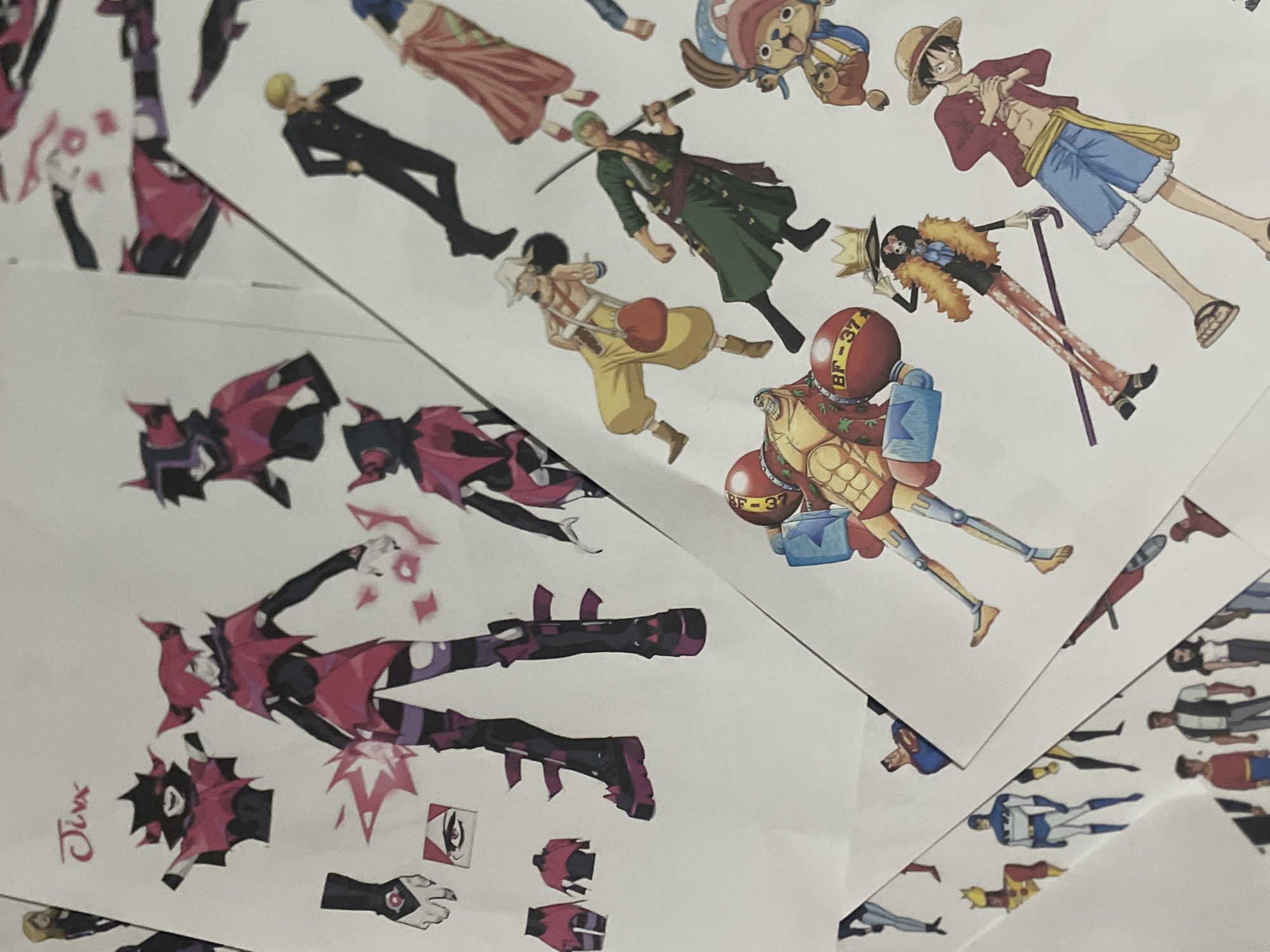 The image is a close-up, overhead shot of several sheets of paper featuring character concept art and stickers. The papers are layered over each other at various angles, obscuring some of the artwork.
The most prominent sheet on the left features concept art of a character named "Jinx," with multiple poses and design elements including her outfit, horns, and magical effects. To the right of this, other papers show characters from the anime series "One Piece," including Monkey D. Luffy, Roronoa Zoro, Usopp, Tony Tony Chopper, and Brook. There are also depictions of characters from DC Comics, such as Superman and Batman, and potentially other superhero characters.
The lighting suggests an indoor setting, likely a desk or table. The focus is sharp on the artwork, with a shallow depth of field blurring the background. There are no discernible elements in the image that would indicate a specific location in Davao City, Philippines.