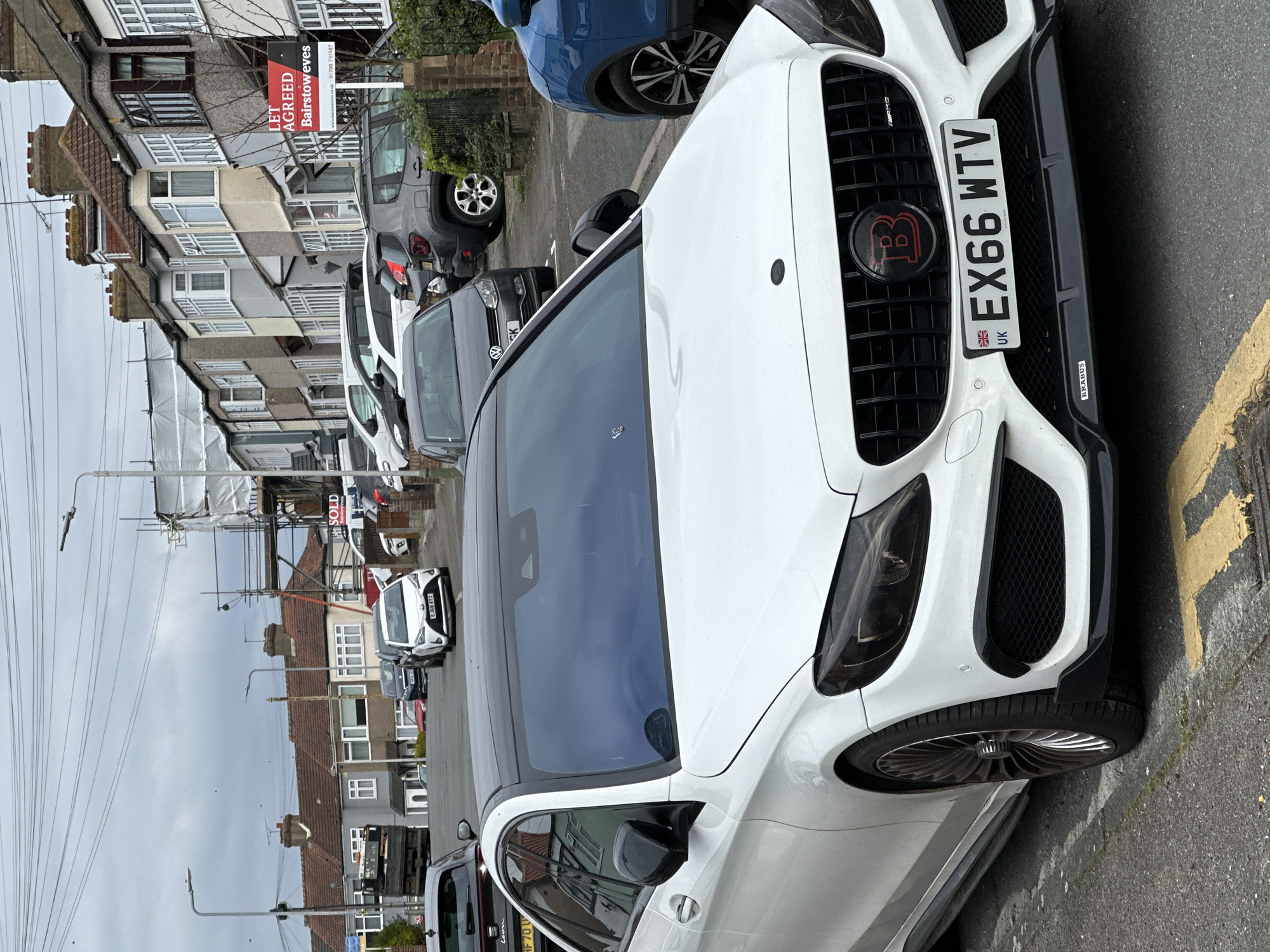 The image depicts a street in what appears to be a residential area in London, United Kingdom. A white Brabus car with license plate "EX66 WTV" is parked on the right side of the street, partially occupying the frame. On the left side of the street, a row of terraced houses is visible, with several other vehicles parked in front of them. A "LET AGREED Bairstoweves" sign is displayed in front of one of the houses, indicating a property transaction. Overhead, utility poles and wires stretch across the street. The sky is overcast, suggesting a cloudy day. The general atmosphere is urban and residential.
