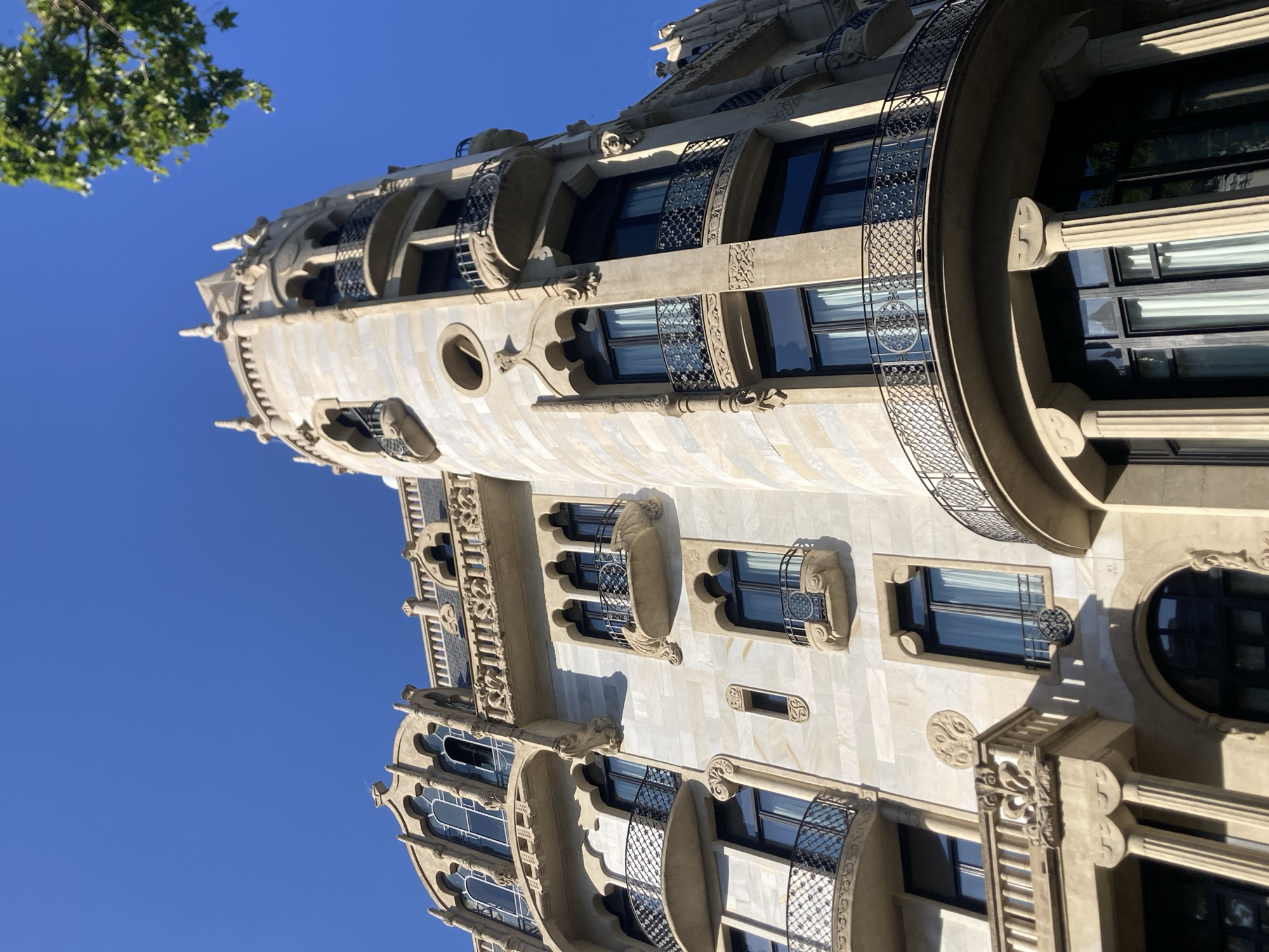 This image captures a low-angle view of a highly ornate Art Nouveau building in Barcelona, Spain, under a clear blue sky. The building's facade is constructed from light-colored stone, possibly marble or limestone, with intricate decorative elements. The architecture features curved lines, elaborate window frames with gothic-inspired tracery, and decorative balconies with wrought-iron railings.
Specific architectural details visible include:
* **Turret:** A prominent cylindrical turret on the right side of the building, topped with decorative spires and featuring several levels of balconies and windows.
* **Windows:** A variety of window styles are present, including large, arched windows with multiple panes on the upper floors and smaller, rectangular windows on lower levels. Many windows are framed with decorative stonework, and some feature small, round decorative elements above them.
* **Balconies:** Several balconies with decorative curved shapes and intricate ironwork are visible, adding to the building's elaborate appearance.
* **Columns:** Large, fluted columns support overhangs and balconies on the lower levels.
* **Decorative Motifs:** The facade is adorned with various decorative motifs, including stylized floral patterns, sculpted figures, and geometric designs.
* **Roofline:** The roofline is characterized by decorative crenellations and spires.
A portion of a leafy green tree branch is visible in the upper right corner, indicating the presence of surrounding foliage. The sunlight casts shadows on the building, highlighting the depth and texture of its architectural features. The scene is devoid of any people or vehicles, focusing solely on the magnificent architecture of the building.
The building's distinctive style and detailed ornamentation strongly suggest it is located in the Eixample district of Barcelona, known for its abundance of Art Nouveau and Modernist architecture. The style is reminiscent of buildings designed by Antoni Gaudí or other prominent architects of that era in Barcelona.