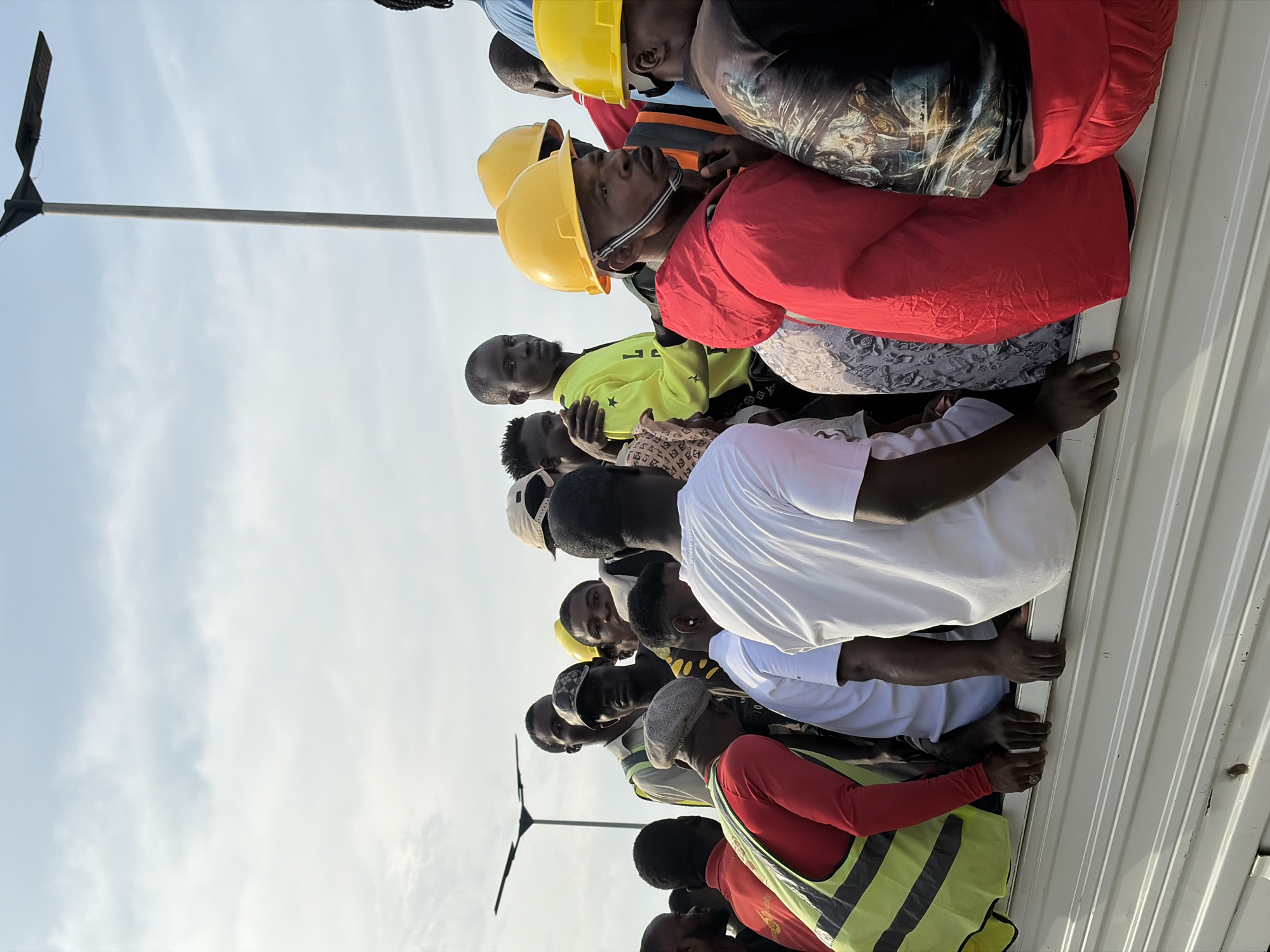 In Alaguntan, Nigeria, a group of men are packed tightly in the back of a white truck. The men are of various ages and are dressed in casual clothing, some wearing hard hats and high-visibility vests, suggesting they are construction workers. The sky above is overcast with a few patches of blue. In the background, a modern street lamp is visible, indicating an urban or semi-urban setting. The truck bed is made of corrugated metal. The overall scene depicts a crowded transportation situation, possibly for laborers.