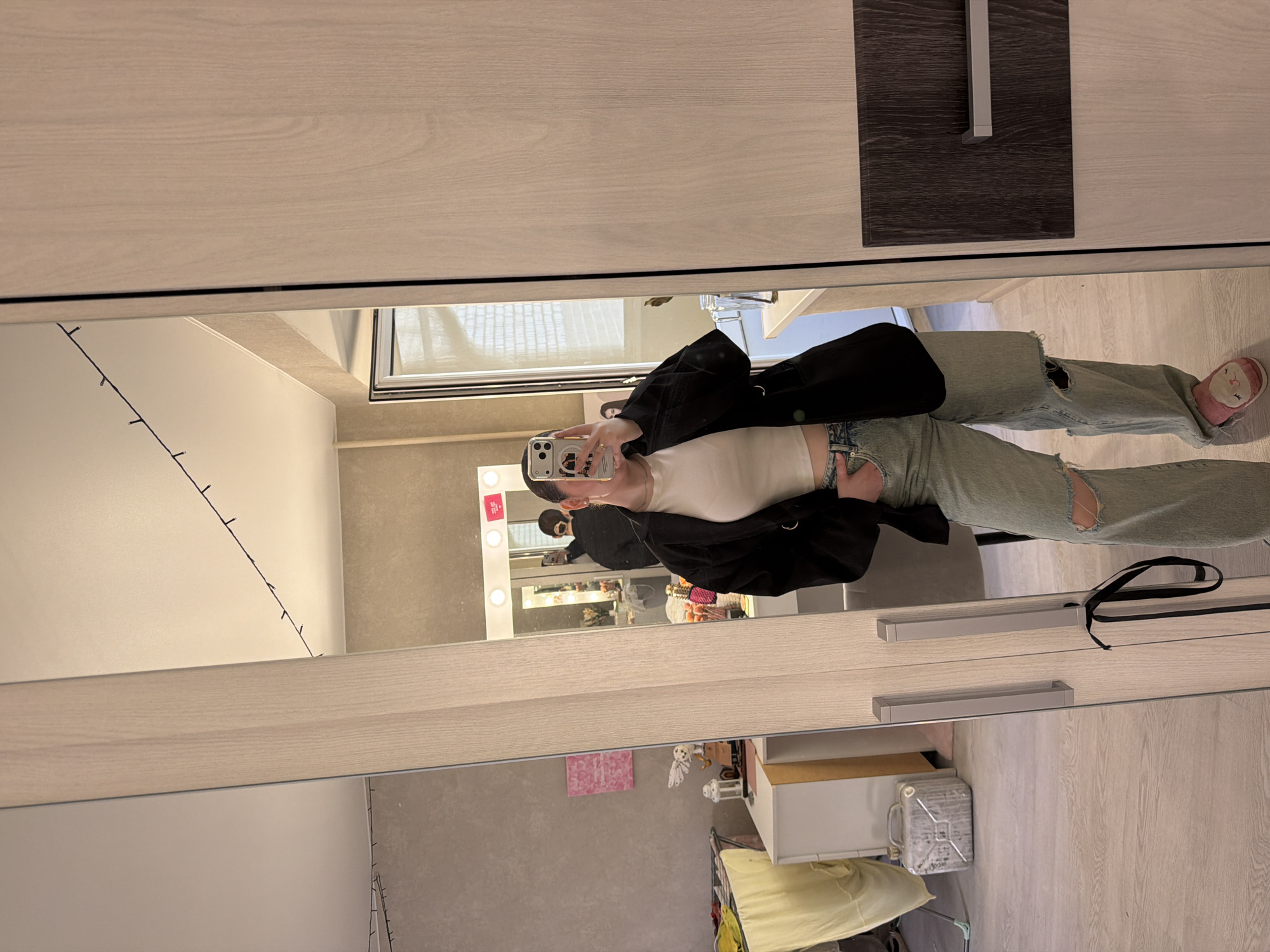 A woman stands in front of a mirror, taking a picture of herself with her phone. She is wearing a white top, ripped light-wash jeans, and a black blazer. Her surroundings include a room with a wardrobe. The wardrobe has light wooden panels and a dark gray rectangular door pull on one side. The room is lit by string lights and a vanity mirror with light bulbs around its perimeter. The reflection in the mirror suggests a dressing area with various beauty and cosmetic items. The location appears to be an interior space, potentially a bedroom or dressing room. Based on the location, this scene might be in the city of Chaikovsky, Russia.