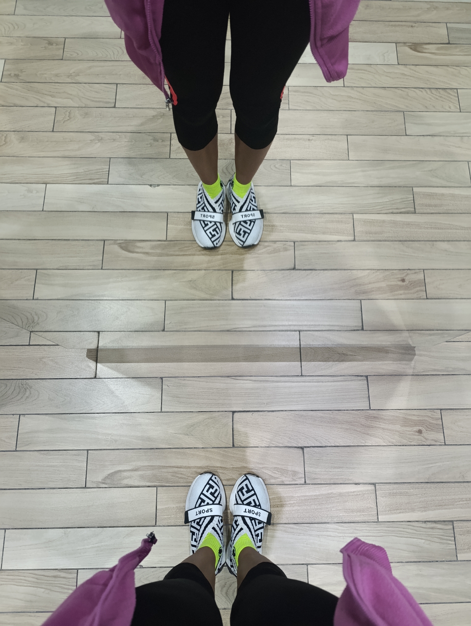 This is a low-angle, overhead shot of two pairs of feet wearing black capri leggings, neon yellow socks, and white sneakers with a black geometric pattern and a black strap that says "SPORT". The feet are standing on a light brown wooden floor with a tiled pattern. The top pair of feet are closer to the camera and have a purple garment draped over the left ankle. The bottom pair of feet are further away and also have a purple garment draped over the left ankle. The scene is indoors, and there are shadows cast on the floor, suggesting artificial lighting. There is no indication of the city of Moscow, Russia.