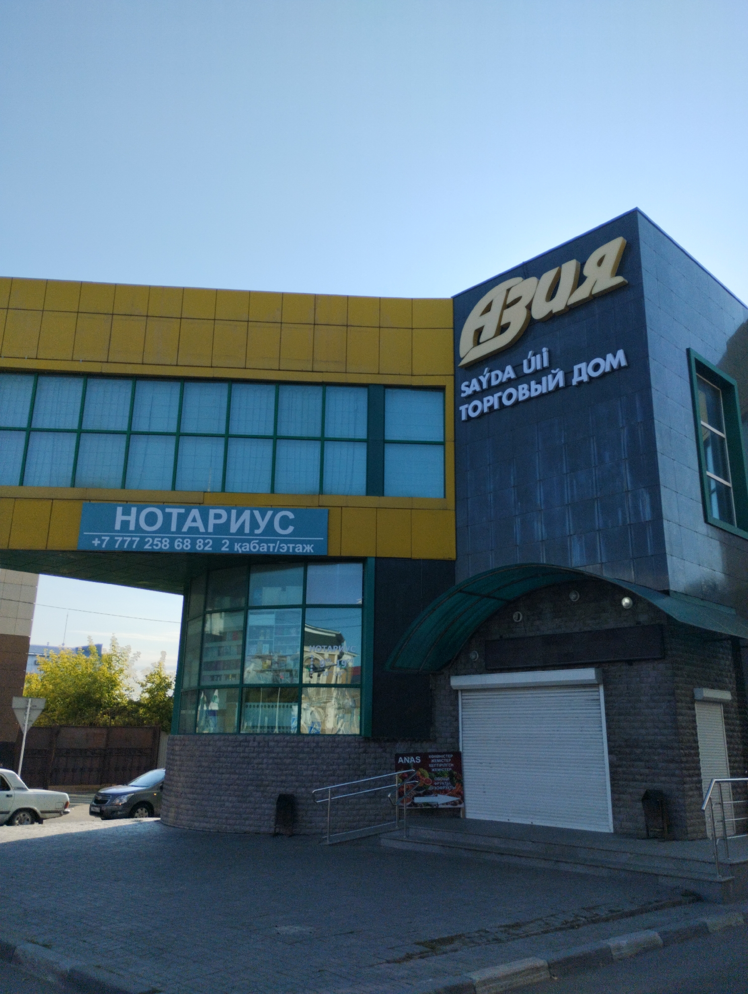The image shows the exterior of a modern building with a prominent yellow upper section and a dark blue tiled lower section. The building has large windows with green frames. A blue sign above a set of windows reads "НОТАРИУС" (Notary) with a phone number and floor information below it. On the dark blue section, a large, stylized Cyrillic word in yellow appears to be a company name, followed by smaller text in English and Cyrillic, possibly indicating a "TRADE HOUSE." A green awning extends over a partially visible storefront with a white roller door. To the left of the building, a light-colored sedan is parked on a paved street. To the right, a dark SUV is also parked. In the foreground, a gray paved sidewalk with a curb is visible. The sky above is clear and blue. A small sign with "ANAS" and images of vegetables and fruits is attached to a metal railing near the storefront. The scene is likely in a commercial area within Kostanay, Kazakhstan.