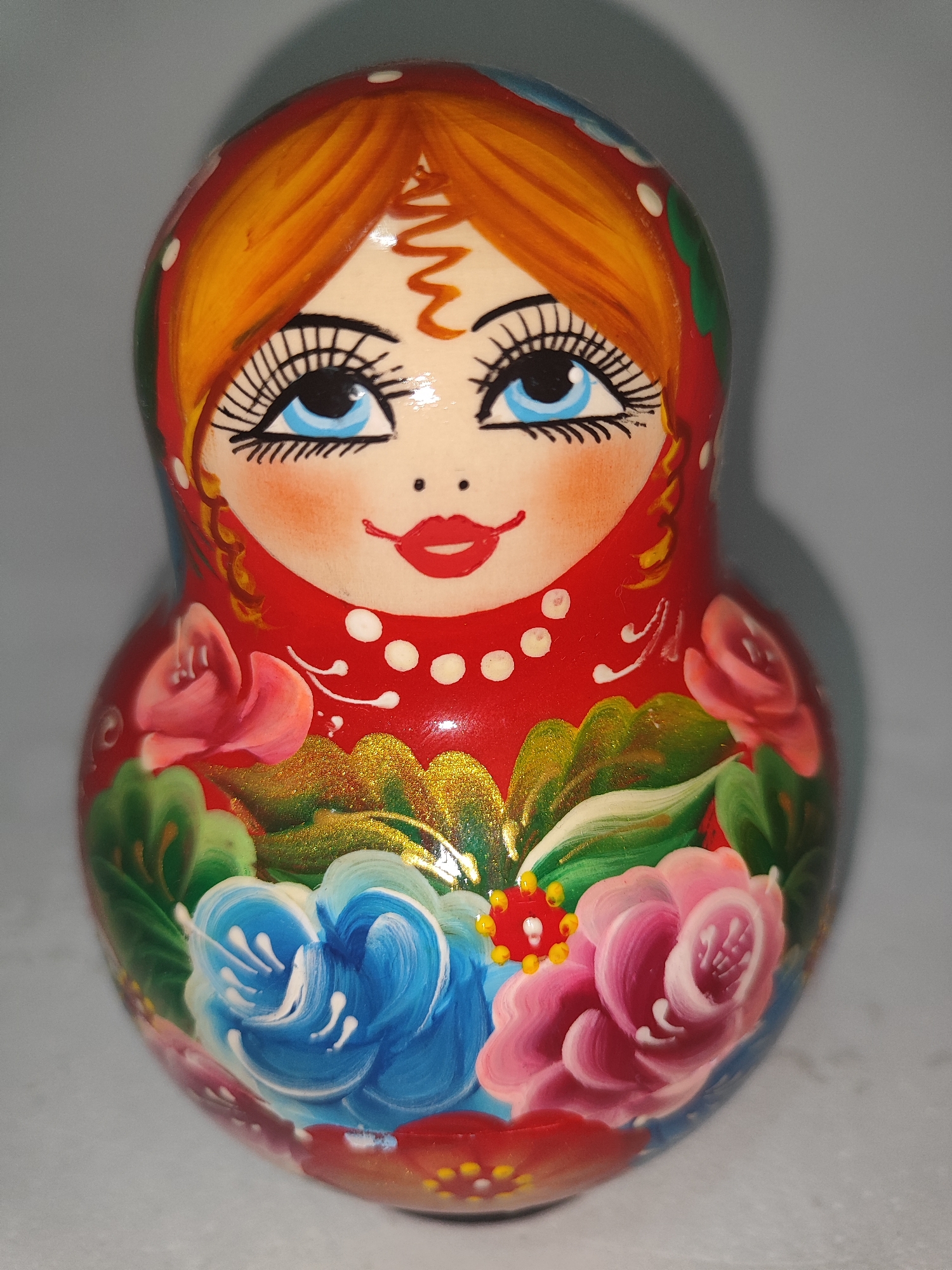 A Russian nesting doll, also known as a Matryoshka doll, is displayed against a plain, light gray background. The doll is painted with a woman's face, featuring blue eyes, rosy cheeks, and red lips. Her hair is a vibrant orange, and she wears a red headscarf adorned with white polka dots and green, red, and blue floral patterns on her lower body. The doll is brightly colored and appears to be made of wood. The lighting casts a subtle shadow to the right of the doll. There are no people or any indication of the doll's specific location within Fergana City, Uzbekistan.
