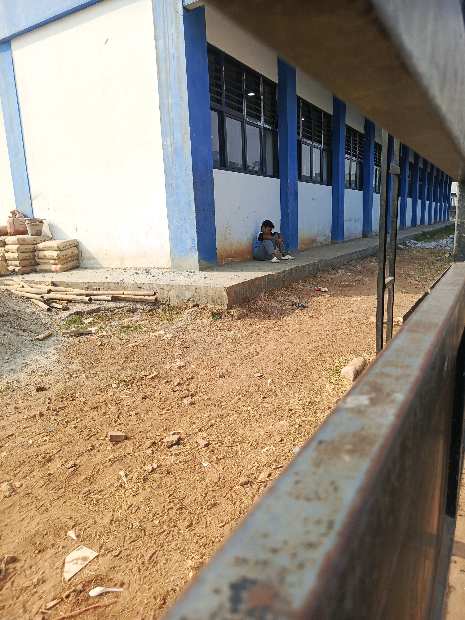 A young boy in a blue shirt and grey pants sits alone against the white wall of a building, his knees drawn up to his chest. He is positioned on a concrete walkway in front of a row of windows with black frames and horizontal louvers. The building's exterior features prominent blue vertical beams. To the left of the boy, several stacks of bags, likely containing cement, are visible, along with a bucket. Bamboo poles and other construction debris are scattered on the ground in front of the walkway. The ground is composed of dry, sandy soil with sparse dry grass and small rocks. A rusty metal fence runs along the right side of the frame, partially obscuring the view. The scene appears to be an outdoor area at a construction site or an unfinished building in Jati Mulya, Indonesia, under daylight.