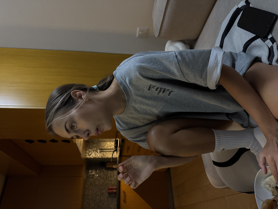 A woman with blonde-streaked hair, wearing a grey t-shirt with text, is squatting. Her gaze is directed downwards, seemingly at something in the kitchen area. She is wearing white socks. A plate with food sits near her hand, close to a couch with a blanket and a phone. The kitchen has orange cabinets, a tiled backsplash, and a faucet with a spout. The lighting is soft, casting shadows in the room.