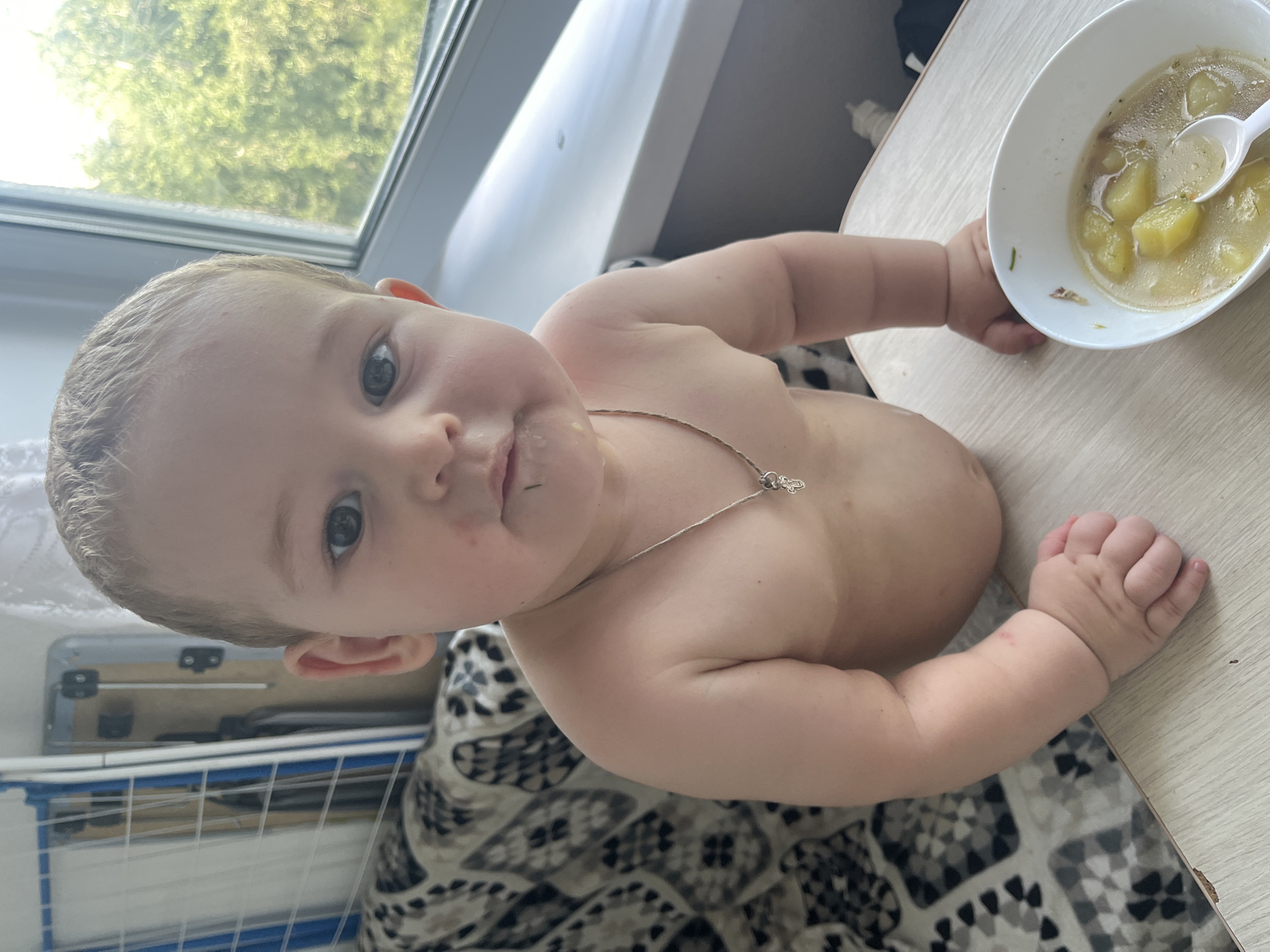 A young child, likely a toddler, is shown from the chest up, seated and facing the camera. The child has light-colored hair and is not wearing a shirt. A silver necklace with a cross pendant is visible around their neck. Their mouth has some food residue around it. To the right of the child, a white bowl filled with soup containing potato chunks and herbs is present on a wooden surface, with a white spoon resting inside. In the background, a patterned cloth is visible on the left, and a window with greenery outside is on the right. The overall lighting suggests an indoor setting during the daytime. The image appears to be taken in an urban residential environment, possibly an apartment, given the presence of a window with a city view and household items like a drying rack in the background on the left. Without further context, pinpointing the exact location within Chelyabinsk, Russia, is not possible.