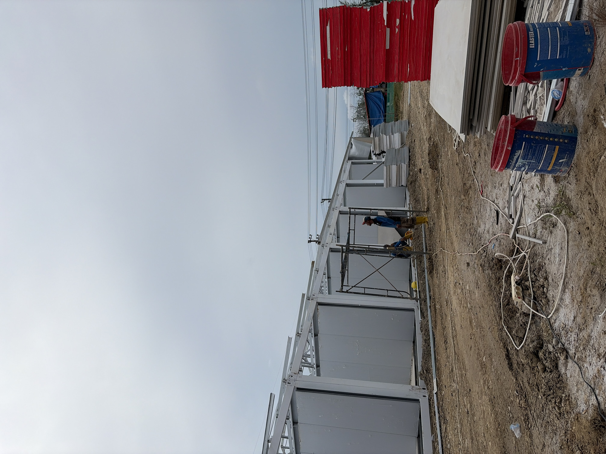 The image depicts a construction site in Simpangempat, Indonesia. A worker is standing on scaffolding, seemingly working on a structure composed of modular, prefabricated units. The structure has a white and grey exterior. To the side are stacks of red and white materials, along with blue buckets labeled "Elastex." The ground is dirt, with visible electrical cords and tools scattered. The sky is overcast.