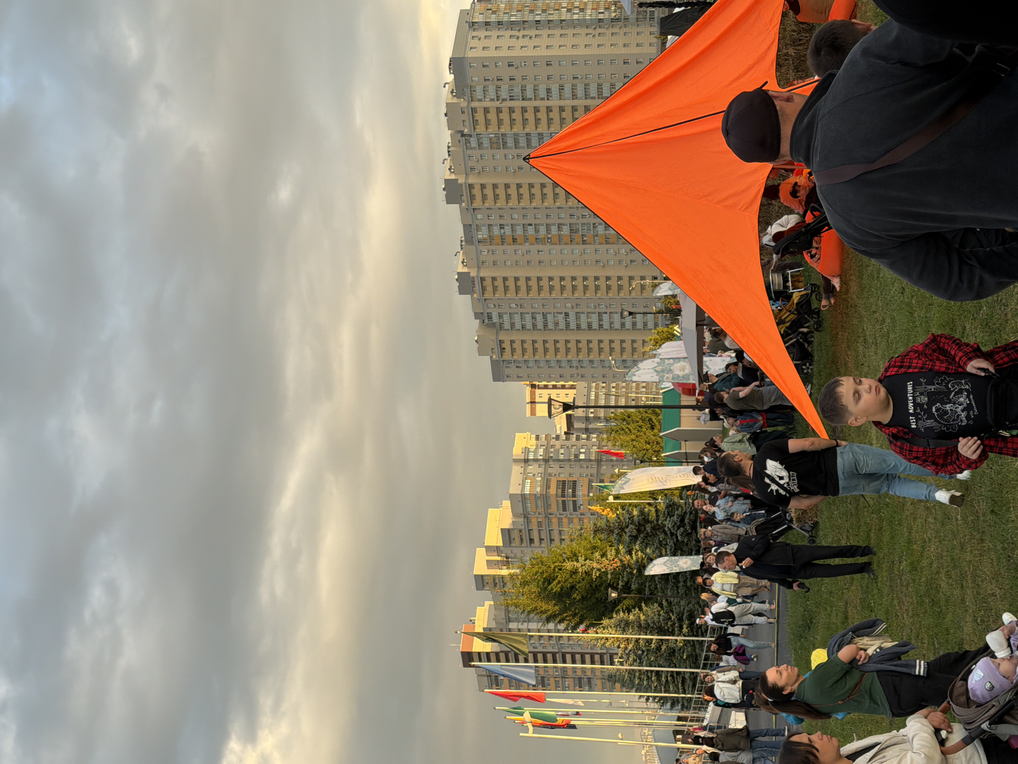 An outdoor event is taking place in an urban setting with apartment buildings in the background. The sky is overcast with patches of lighter clouds, and the setting sun casts a warm glow on the buildings. In the foreground, a large, bright orange tent dominates the scene, its triangular peak pointing upwards. A crowd of people are milling around on a grassy area. Some are standing and talking, while others are seated or pushing strollers. A young boy in a red and black plaid shirt is visible in the lower right corner, looking towards the right. A man in a dark jacket and black baseball cap is seen from behind, facing the orange tent. Behind the tent, several people are sitting or lying down. In the mid-ground, a row of evergreen trees stands tall, with several flags on poles planted in front of them. The flags are of various colors and designs, including a blue flag and a green flag. The apartment buildings in the background are modern and appear to be several stories high, with many windows. The overall atmosphere appears to be relaxed and festive.