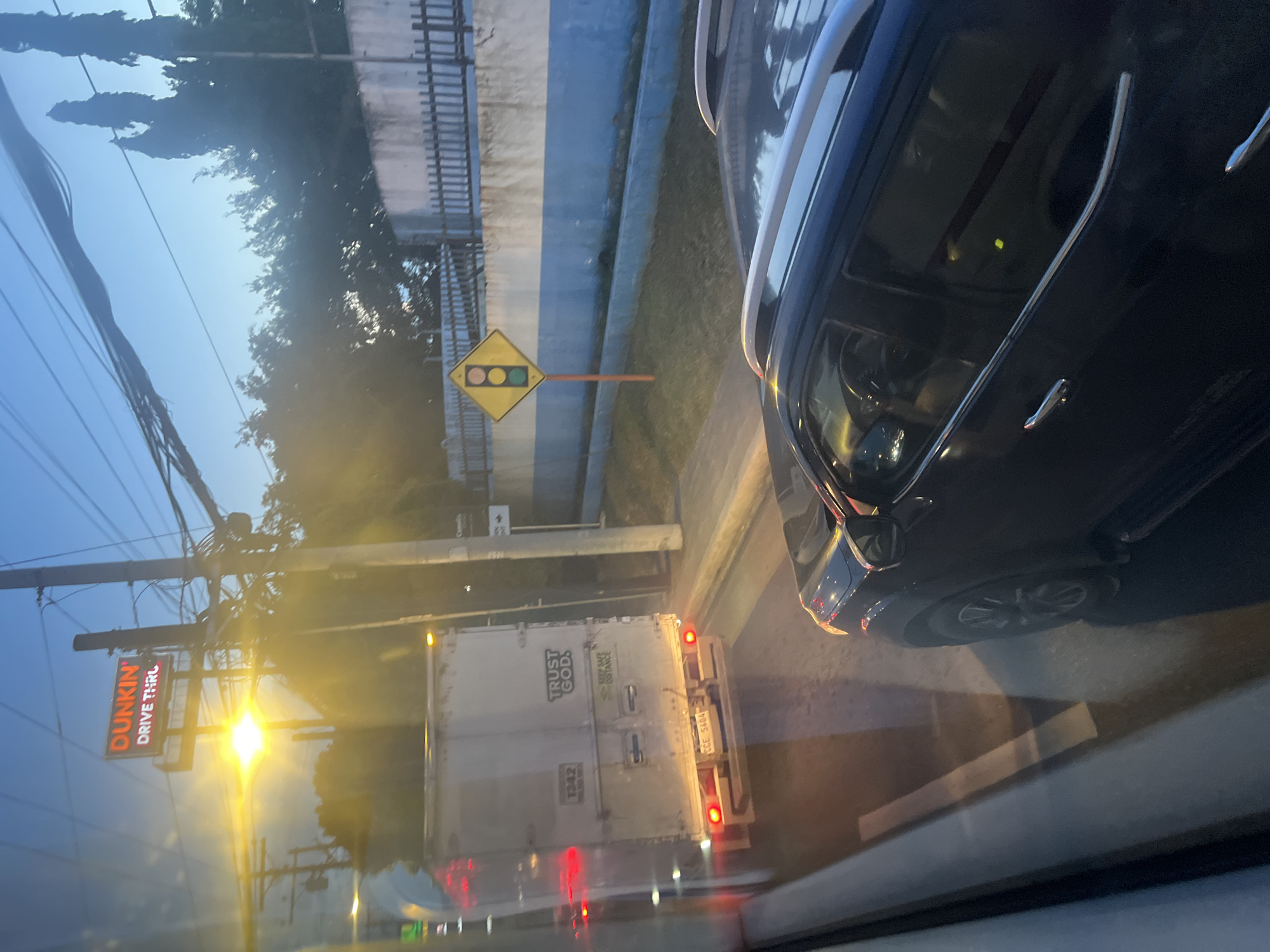 The image captures a traffic scene in Davao City, Philippines, during twilight. In the foreground, the dark silhouette of a vehicle is visible. Further ahead, a white truck with its hazard lights illuminated is on the road, with the license plate "CCE 5404" partially visible. The truck displays the text "TRUST GOD." and "KEEP SAFE DISTANCE." A large, illuminated "DUNKIN' DRIVE THRU" sign is prominently displayed above the road. A traffic signal, showing a yellow light, is positioned on a pole. Utility poles with numerous wires crisscross the sky. The environment suggests an urban setting with trees and a concrete fence visible in the background. The overall lighting indicates it is either dawn or dusk.