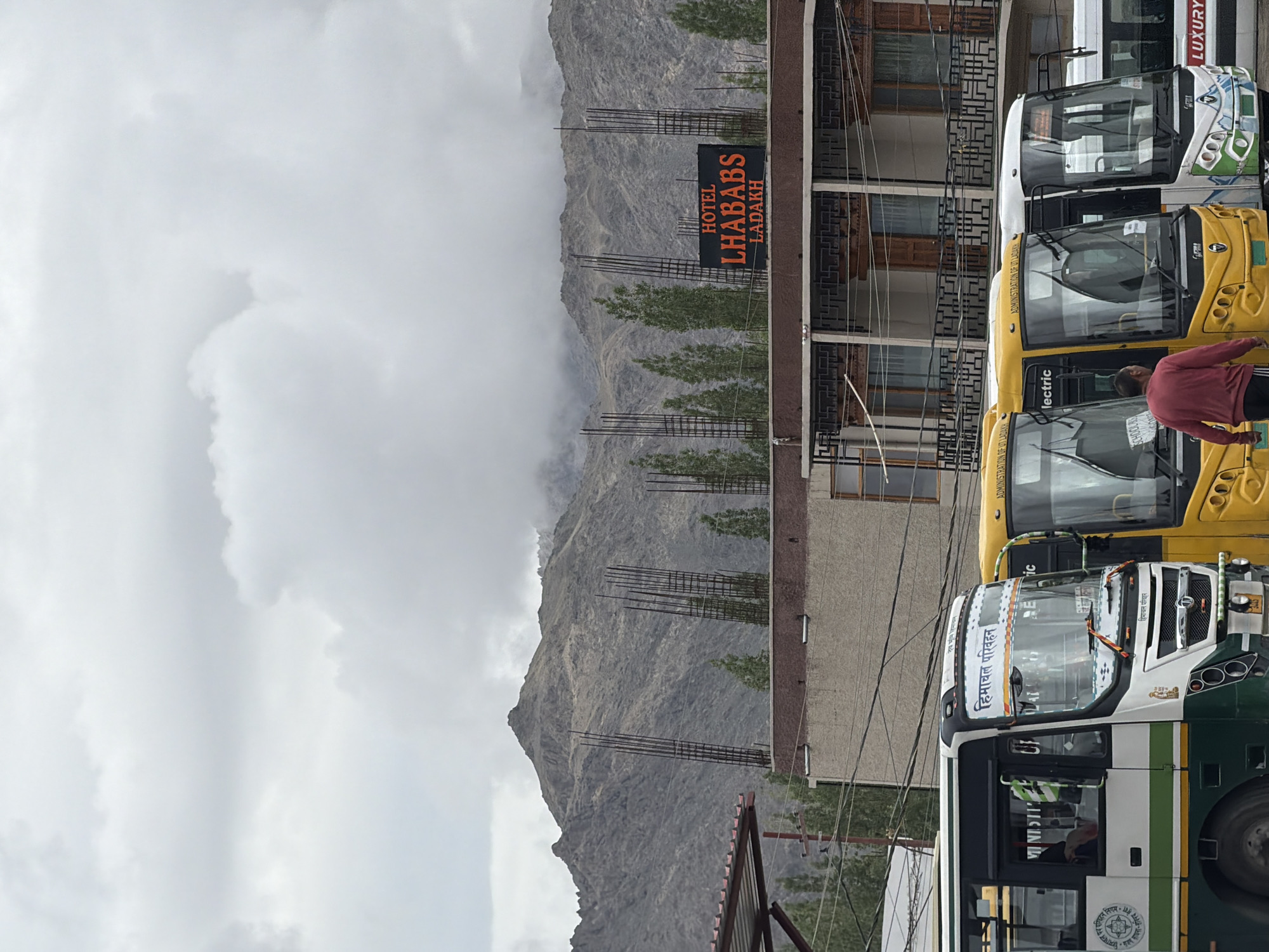 The image depicts a street scene in Leh, Ladakh, India, under a cloudy sky. In the foreground, several buses are parked. The bus on the left is white and green with "HIMACHAL PARIVAHAN" written in Hindi on its windshield. To its right are yellow buses, one of which has "ADMINISTRATION OF UT LADAKH" written on its windshield. A person in a red shirt is walking away from the viewer towards the yellow buses. Behind the buses is a building with a sign that reads "HOTEL LHABABS LADAKH." Further in the background, a mountain range is visible, partially obscured by clouds. The environment suggests a somewhat overcast day in a mountainous region.