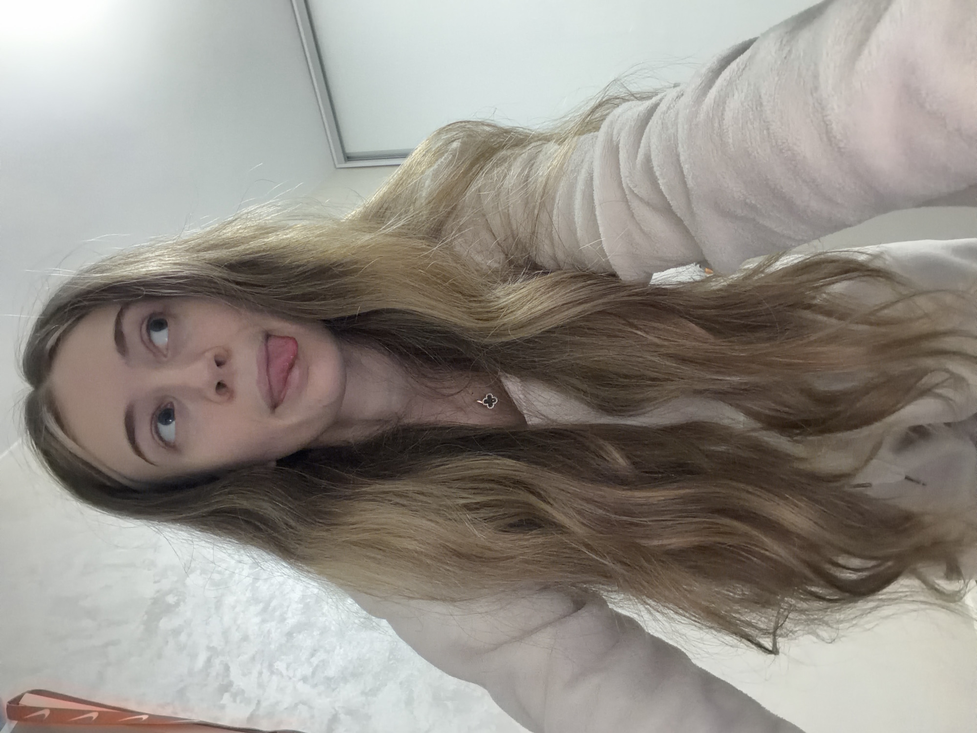 The image shows a woman with long, wavy, highlighted blonde and brown hair sticking out her tongue. She is wearing a light-colored, possibly white or beige, fluffy robe or sweater. A small, clover-shaped necklace is visible around her neck. The background is blurry, but it appears to be an interior setting with a white wall or surface behind her. It is impossible to determine if the picture was taken in Plovdiv, Bulgaria.