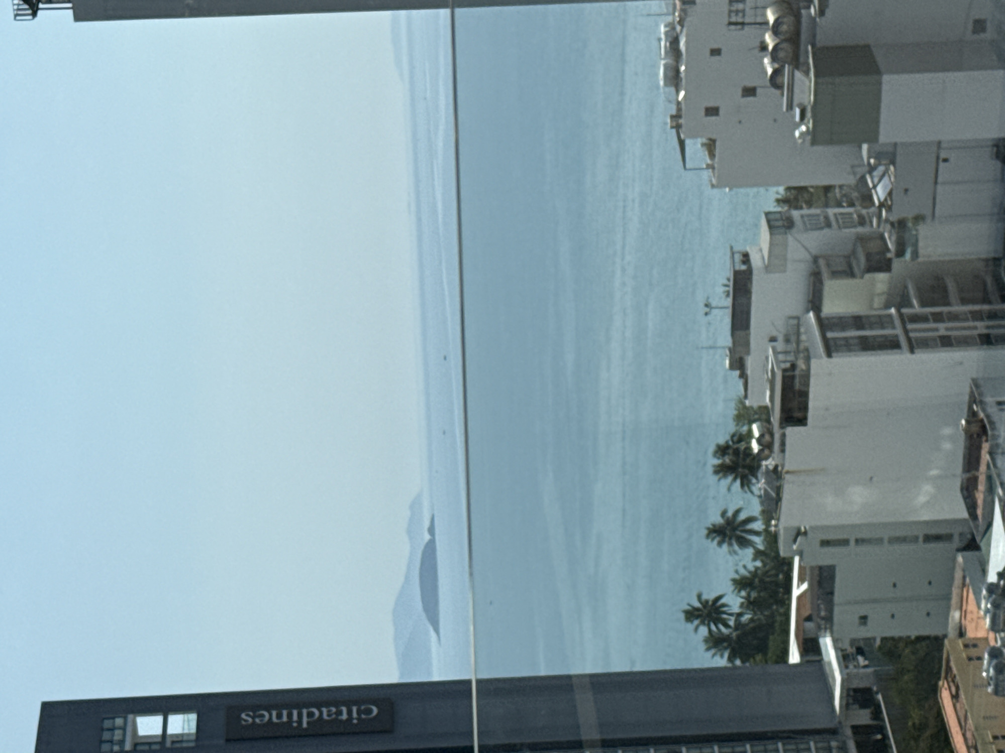 The image shows a view of the ocean and coastline in Nha Trang Ward, Vietnam, as seen from a high vantage point. The "citadines" building is visible in the foreground. The ocean is a light blue color, and the coastline is lined with buildings and palm trees. The buildings are mostly white and gray, and they vary in height. The sky is clear.